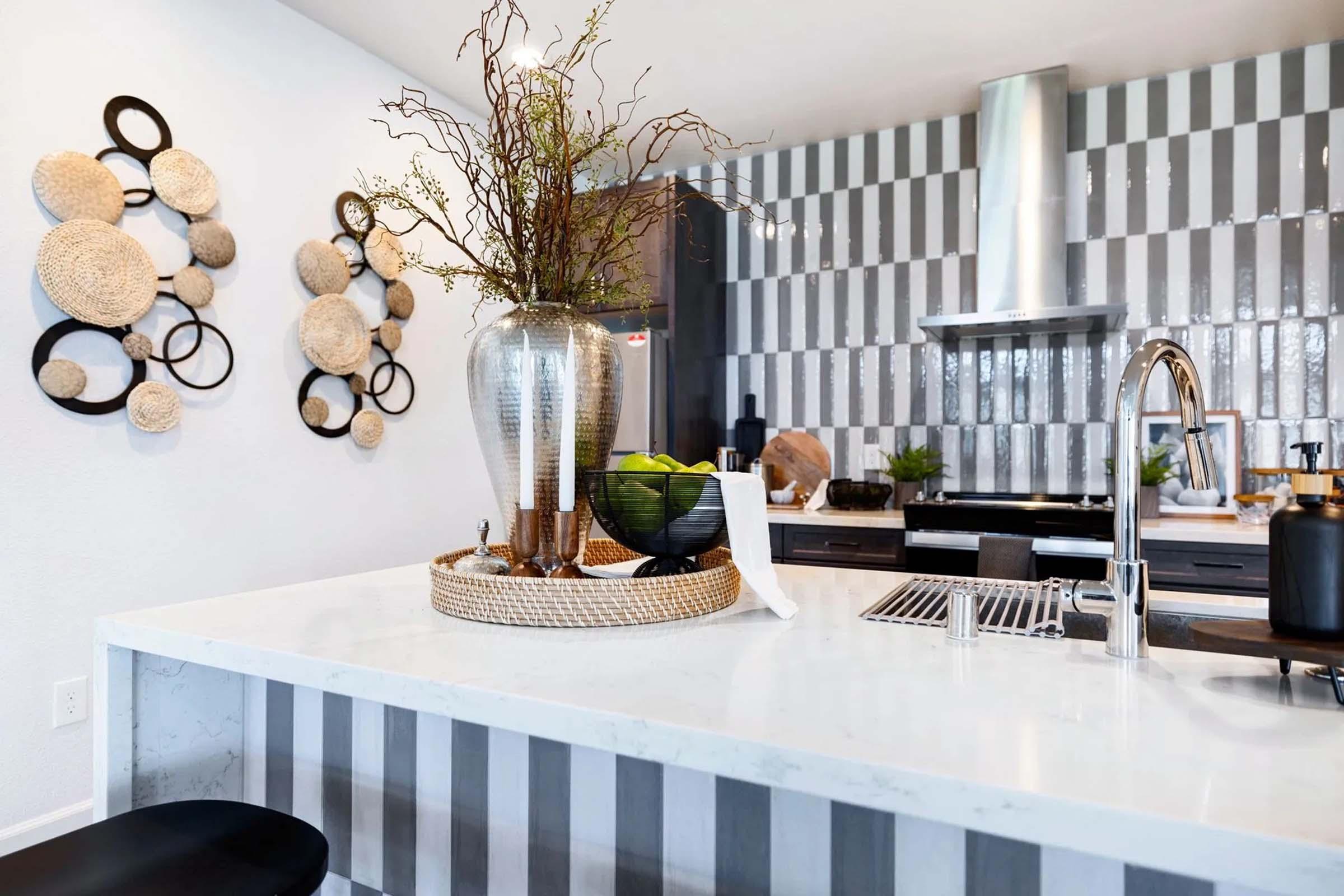 A modern kitchen featuring a marble countertop, stylish decor with decorative baskets on the wall, a sleek stainless steel range hood, and a black bowl filled with green fruit. The backsplash showcases a contemporary design with varying textures, creating an inviting atmosphere.