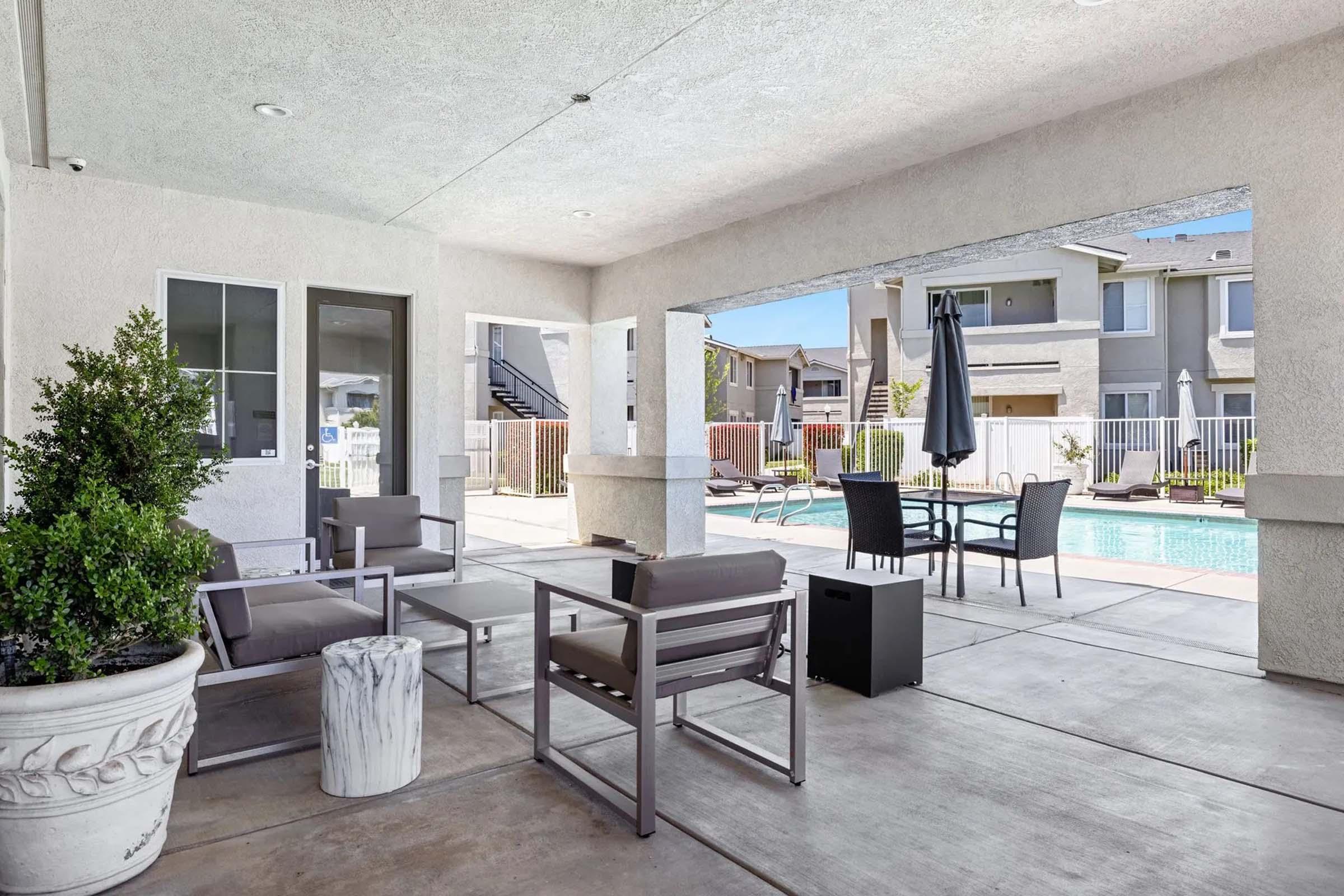 A modern outdoor seating area featuring gray patio furniture, including a table and chairs, under a shaded architectural overhang. In the background, a swimming pool and residential buildings can be seen, surrounded by a fence and landscaped greenery.