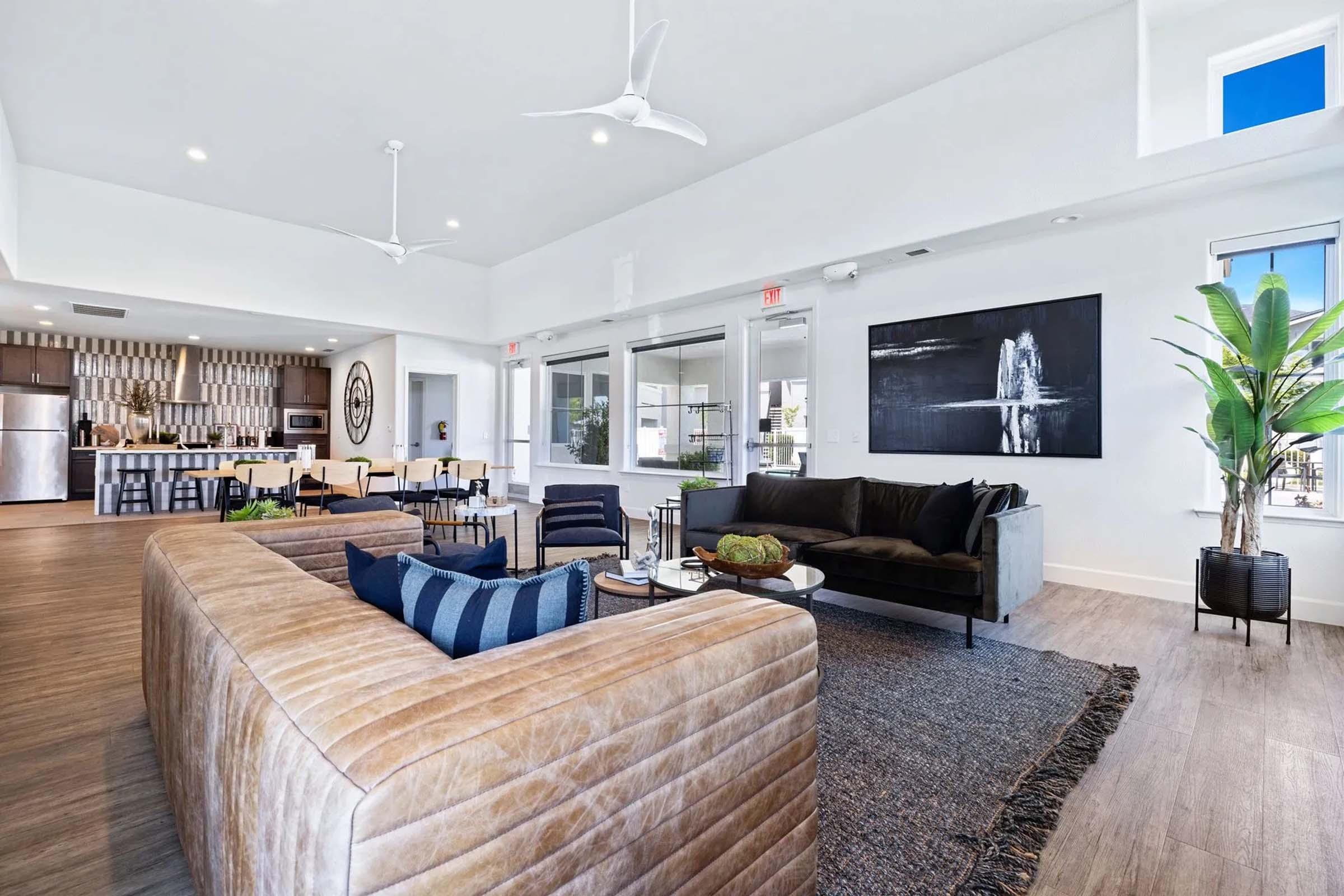 Modern living room featuring a large, plush sofa with textured upholstery, a stylish coffee table, and decorative elements. The space includes a dining area visible in the background, with sleek furniture and a contemporary wall art piece. Large windows allow natural light to fill the room, complemented by greenery.