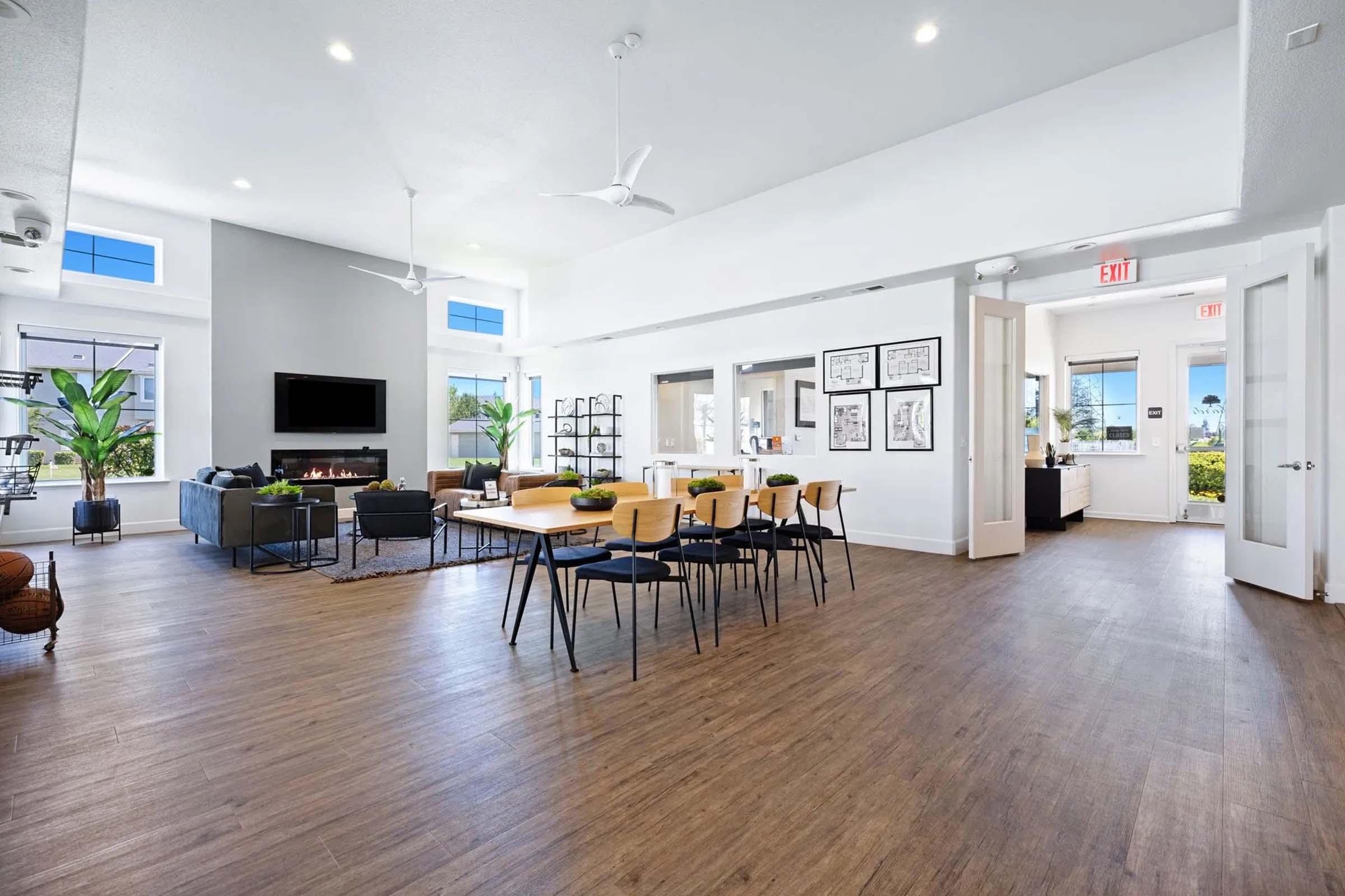 A spacious, modern common area featuring a dining table with chairs, a cozy seating arrangement, large windows allowing natural light, plants for decoration, and stylish artwork on the walls. There is a TV mounted on a wall and open doorways leading to other areas. The flooring is wooden.
