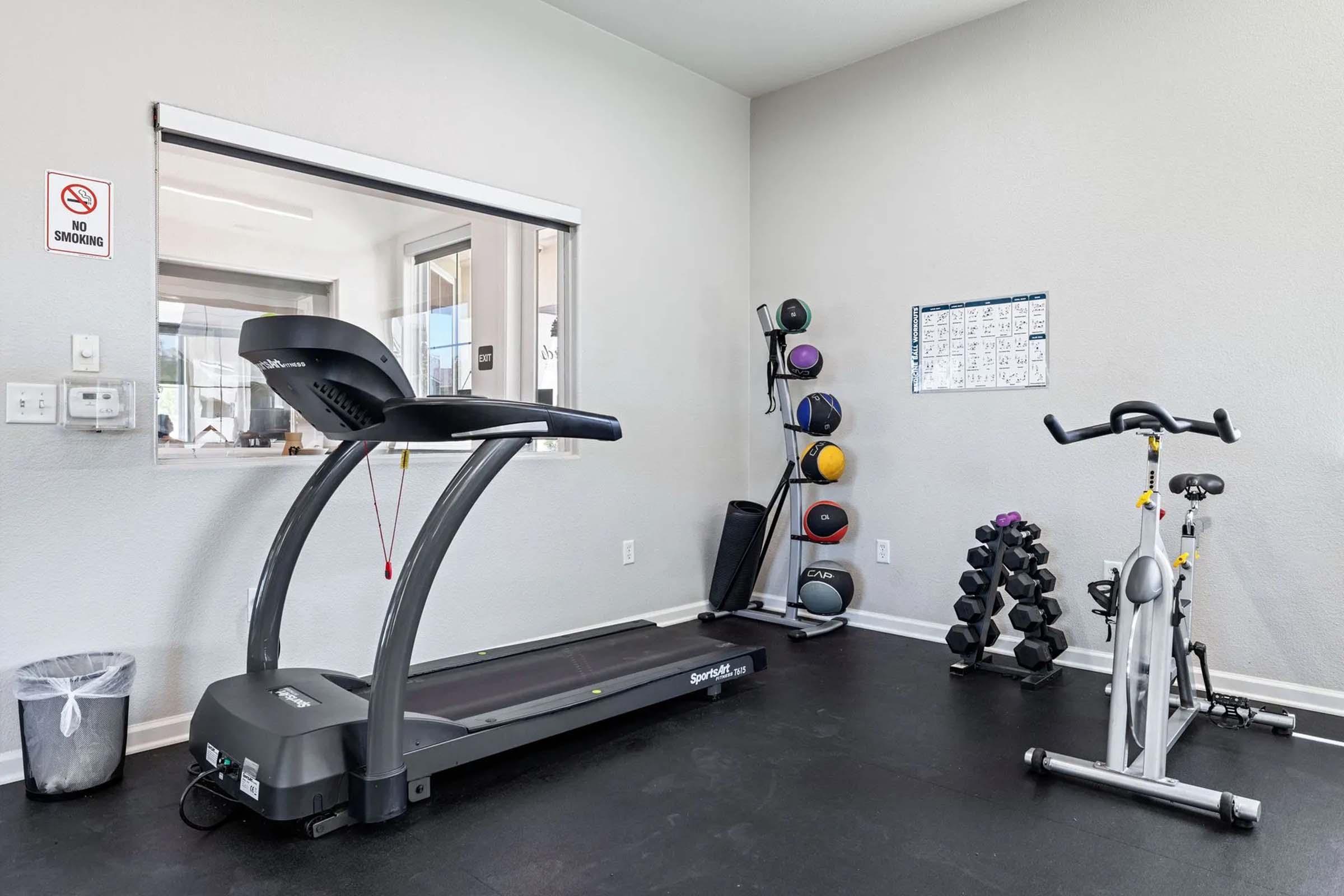 A small gym space featuring a treadmill and an exercise bike, with weights neatly stacked on a rack. There's a no smoking sign on the left wall, and a calendar or schedule is mounted on the wall opposite the equipment, creating a functional and organized workout environment.