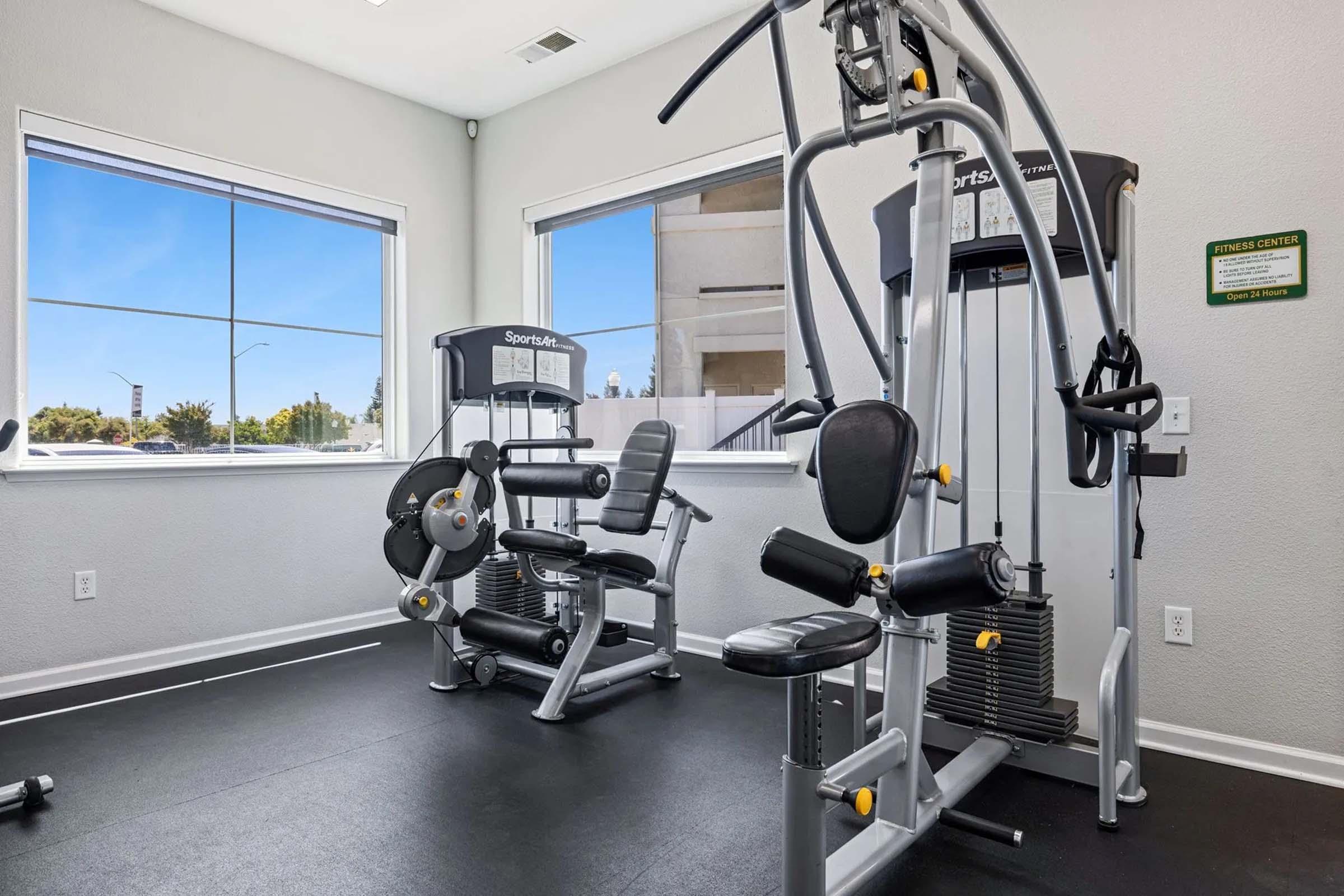 A modern gym interior featuring various exercise equipment, including a multi-station gym machine and a leg press. The room has large windows allowing natural light, creating an inviting atmosphere for workouts. The floor is covered with black mats, and there are motivational signs on the walls.