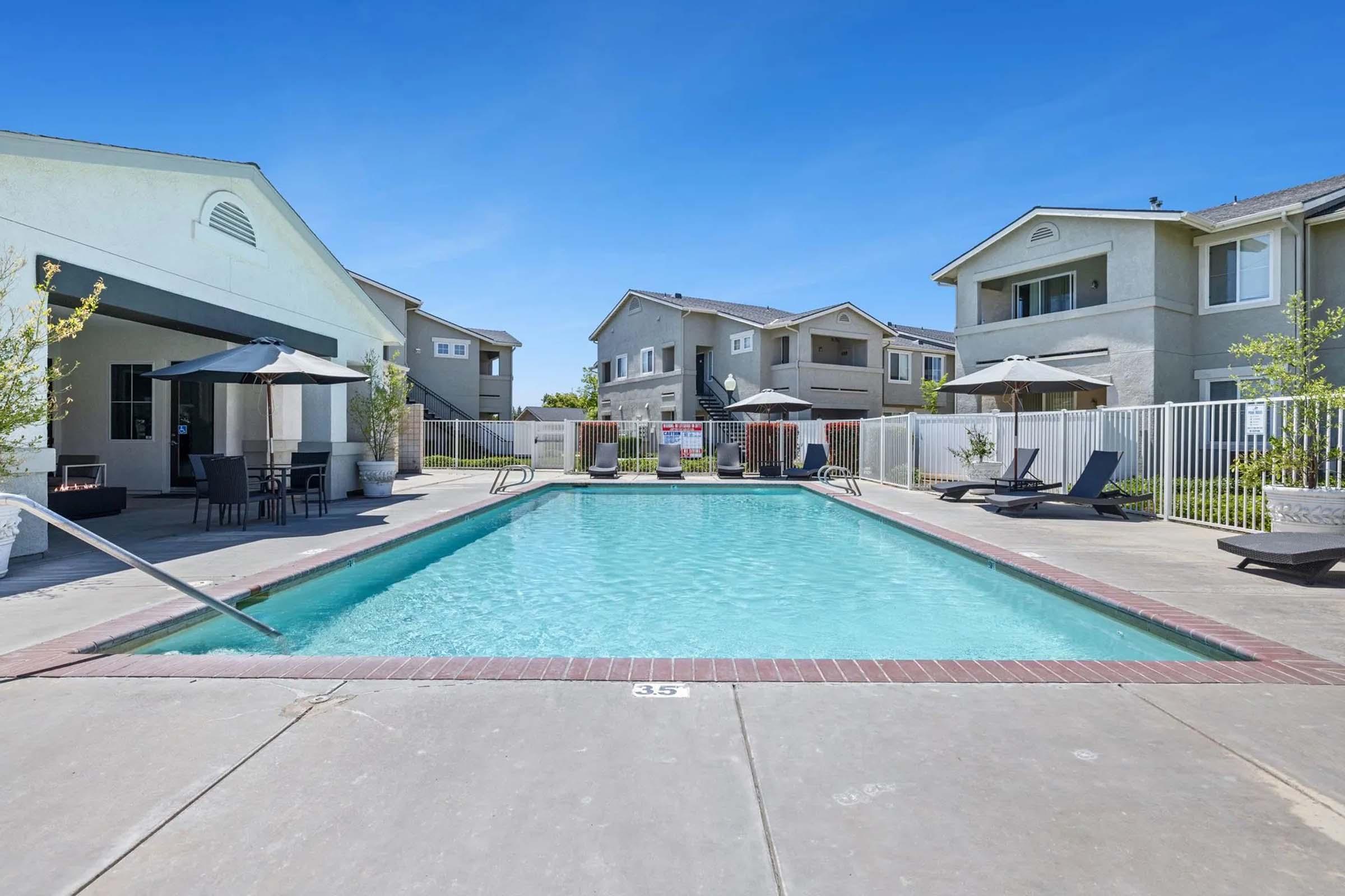 A swimming pool surrounded by lounge chairs and umbrellas, situated within a residential complex. The clear blue water reflects the blue sky, and nearby, there are two-story buildings with balconies. A white fence encloses the pool area, creating a relaxing outdoor atmosphere.