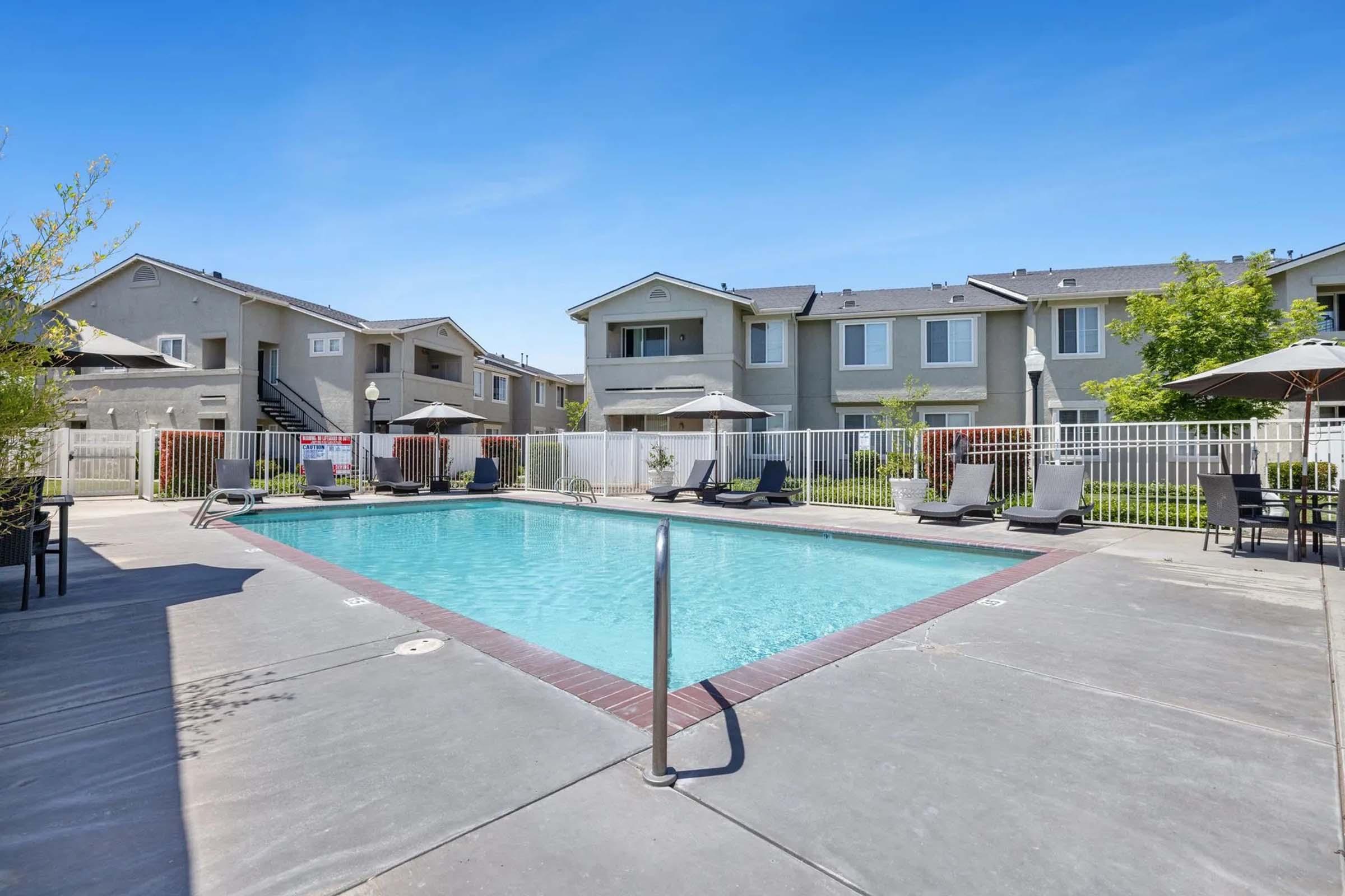 A clear outdoor swimming pool surrounded by lounge chairs and patio umbrellas, with a residential building in the background. Bright blue sky and greenery create a welcoming atmosphere.