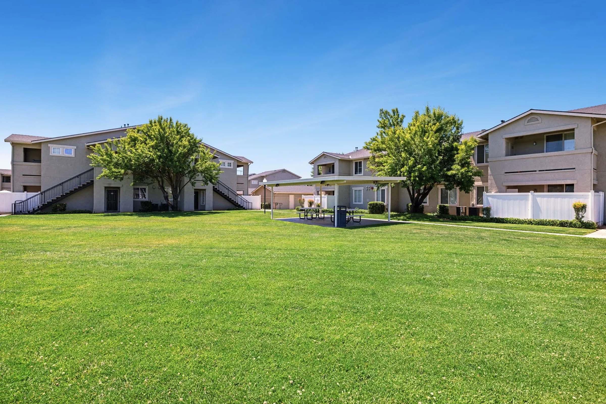 A spacious outdoor area featuring well-maintained green grass, surrounded by residential buildings. There are trees providing shade and a small gazebo with tables and chairs, perfect for relaxation or social gatherings. The sky is clear and blue, creating a pleasant atmosphere.