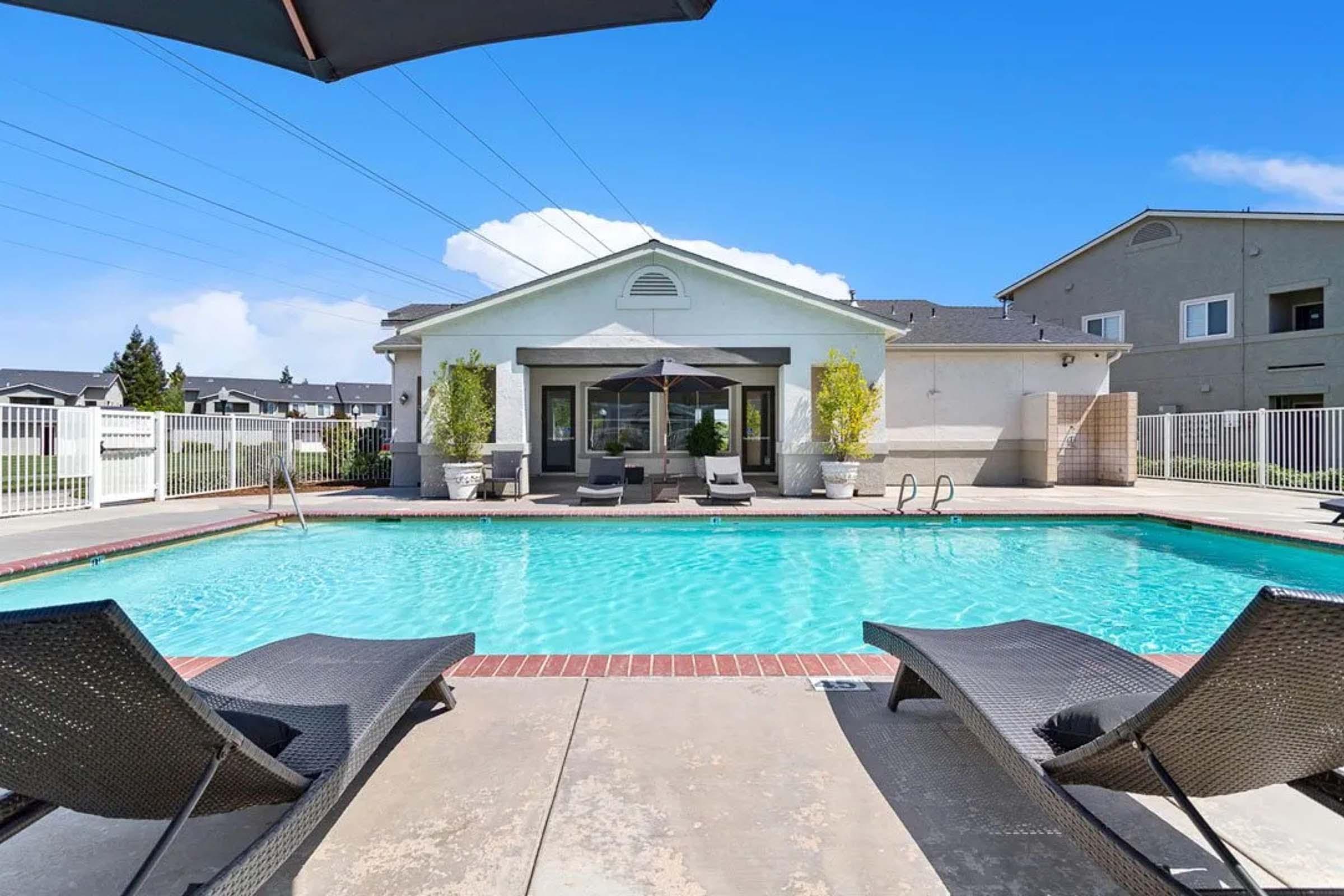 A serene outdoor pool area with lounge chairs on the deck, surrounded by a white fence. Behind the pool, there's a modern building featuring large windows and a covered patio. The sky is clear and blue, adding to the inviting atmosphere.