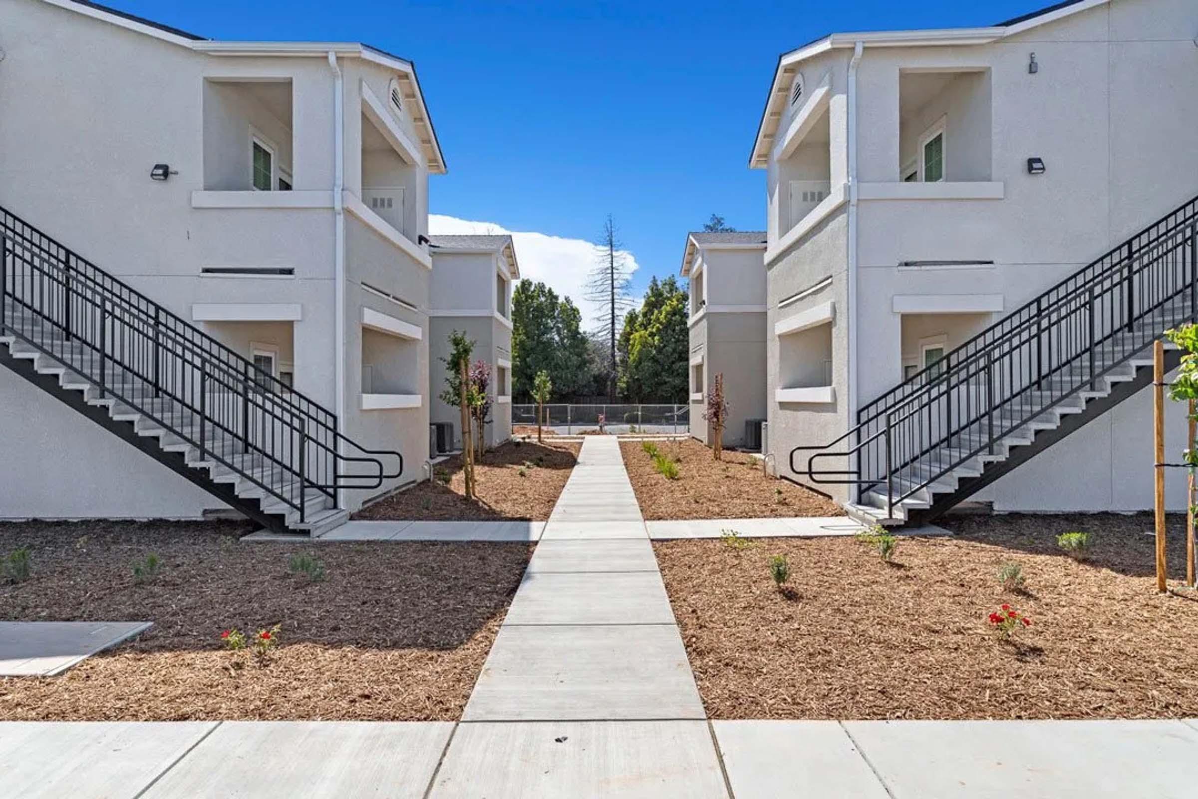 Two identical two-story apartment buildings with staircases on either side, separated by a concrete pathway. The area is landscaped with small plants and flowerbeds. The sky is clear and blue, creating a bright and inviting atmosphere.