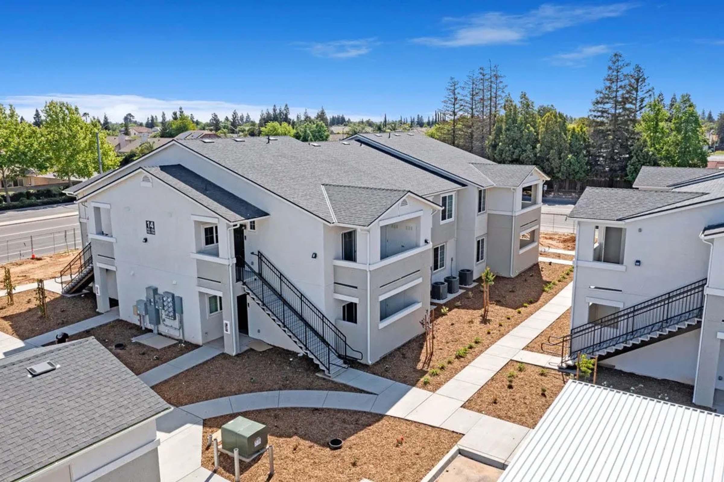 Aerial view of a modern apartment complex featuring two-story buildings with gray roofs. The courtyard has newly landscaped areas with small plants and a pathway. Surrounding trees and a road can be seen in the background, indicating a residential neighborhood.
