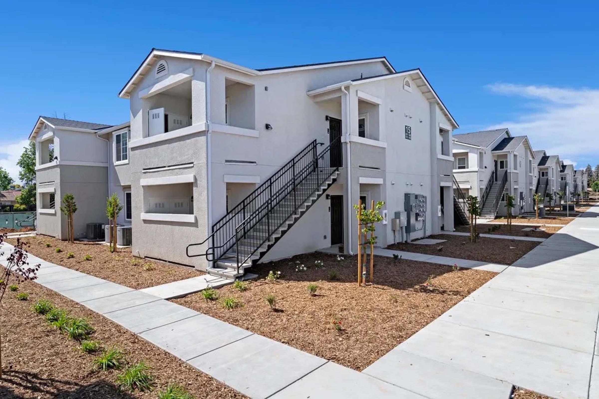 A modern, two-story apartment building with a light-colored exterior, featuring multiple units, staircases, and a landscaped area. The pathway in front is lined with young plants and shrubs, and the sky above is clear and blue.