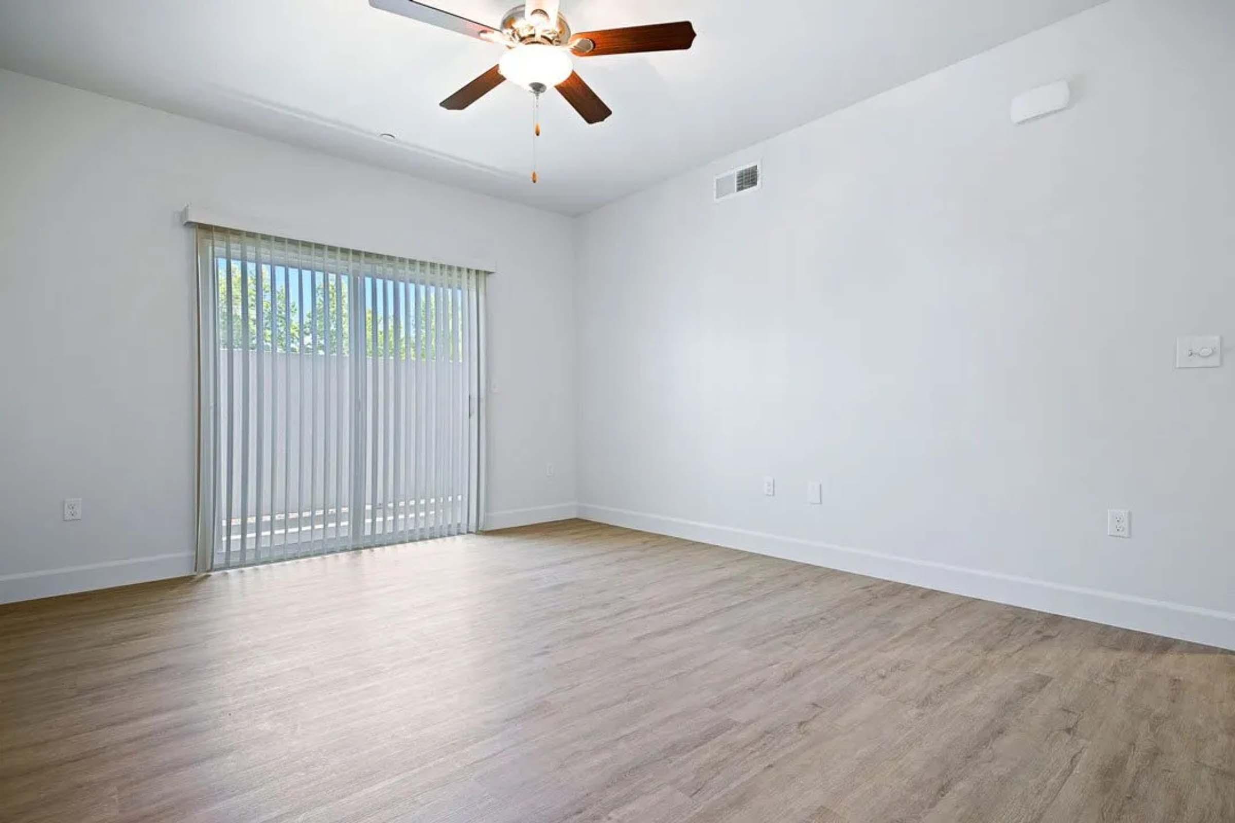 A bright, empty room featuring a ceiling fan, light-colored walls, and wood-like flooring. There is a sliding glass door with vertical blinds leading to an outdoor area, allowing natural light to enhance the spacious, minimalist design.