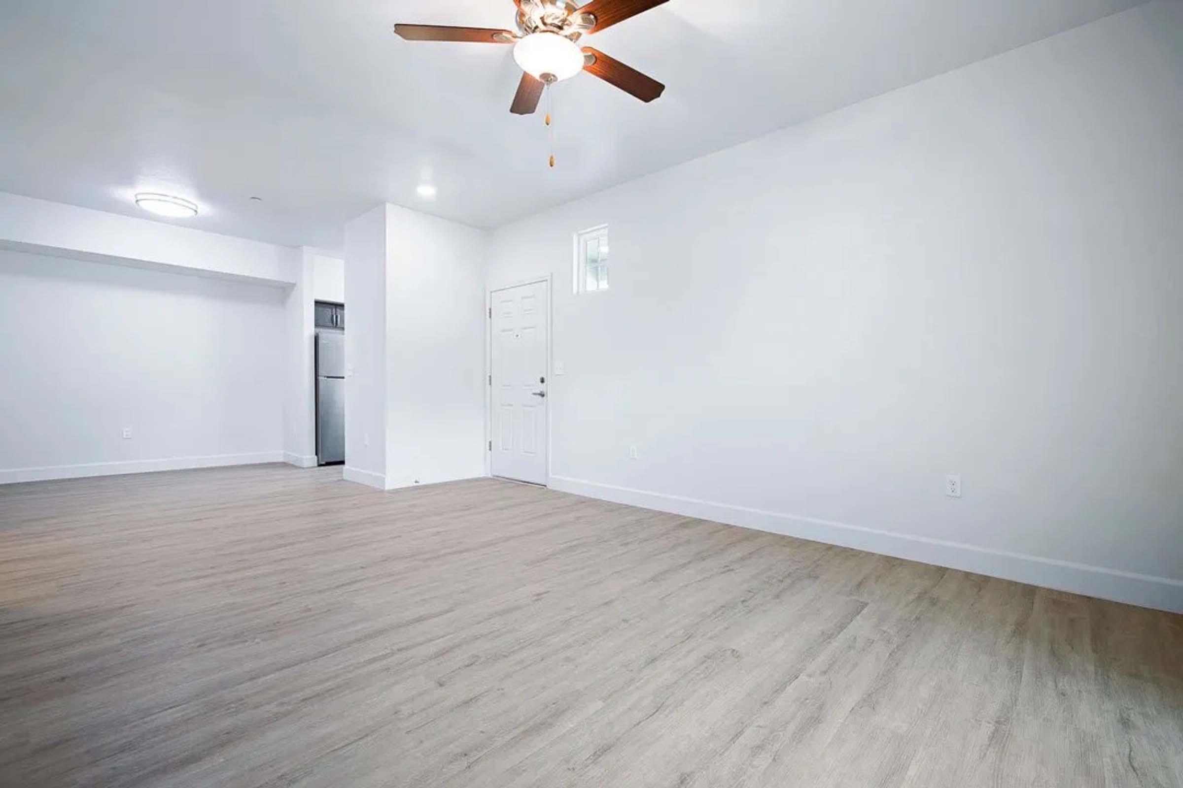 A spacious, empty room with light-colored walls and hardwood flooring. A ceiling fan is visible, and there is a door and a window on one wall. In the background, a kitchen area with stainless steel appliances can be seen, creating a bright, open atmosphere.