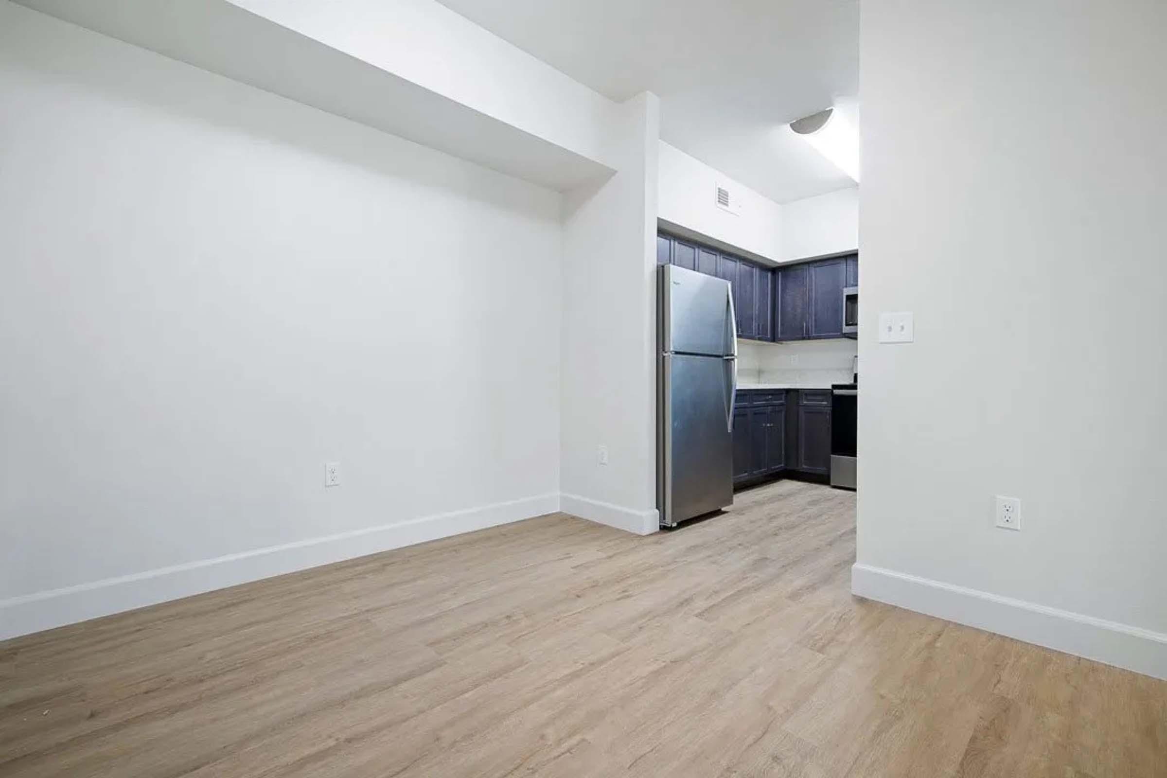 A spacious, modern kitchen area featuring a stainless steel refrigerator, dark wooden cabinets, and light-colored walls. The flooring is light wood, creating a bright and inviting atmosphere.