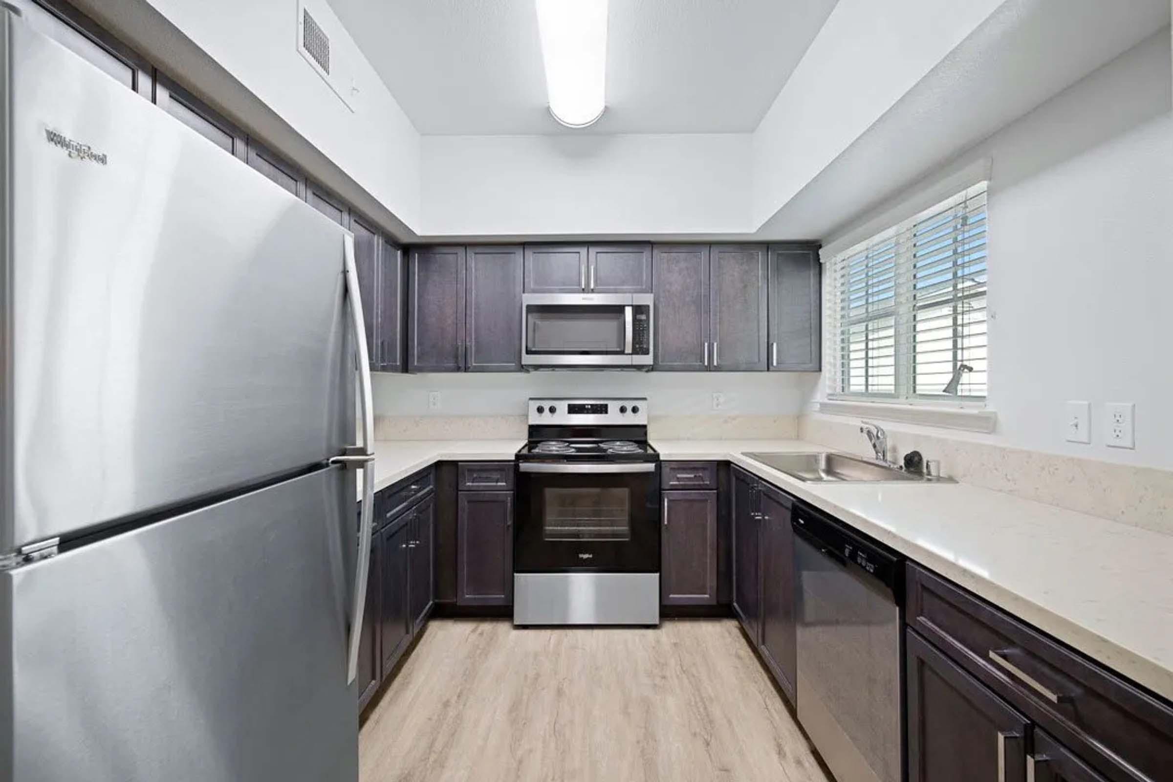 Modern kitchen featuring stainless steel appliances, including a refrigerator, oven, and microwave. Dark wood cabinets contrast with light countertops. A sink is positioned beside a dishwasher. Bright, natural light streams through a window with horizontal blinds. Light-colored flooring completes the contemporary look.