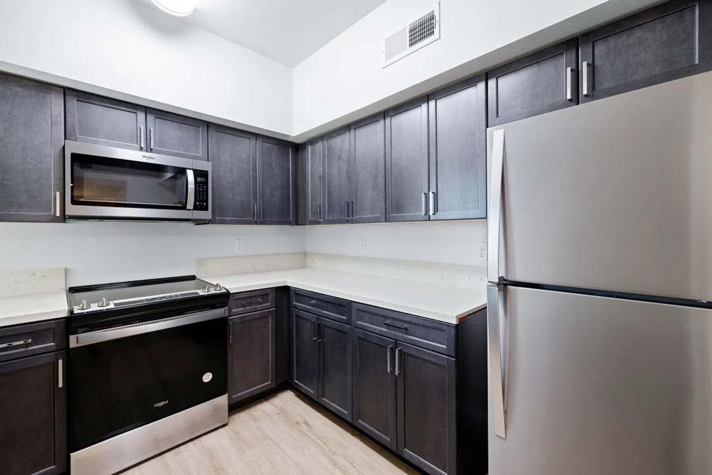 Modern kitchen featuring dark wood cabinets, a stainless steel refrigerator, a built-in microwave above the stove, and a smooth countertop. The space has a neutral color scheme with wood-style flooring, creating a sleek and functional environment.