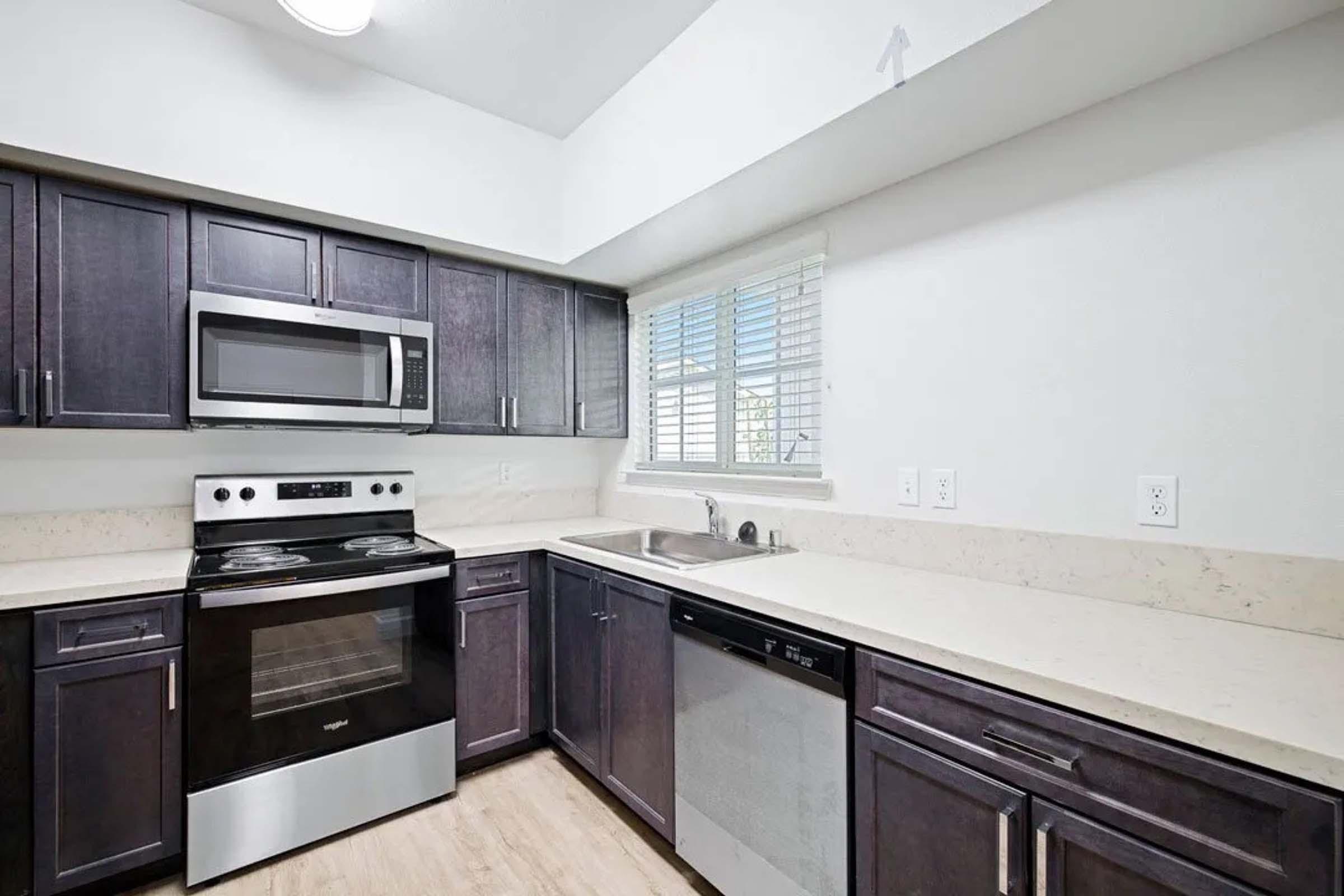 Modern kitchen featuring dark wooden cabinets, stainless steel appliances including an oven, microwave, and dishwasher. Light-colored countertops and a window providing natural light. The layout is spacious with a neutral color palette, creating a clean and contemporary look.