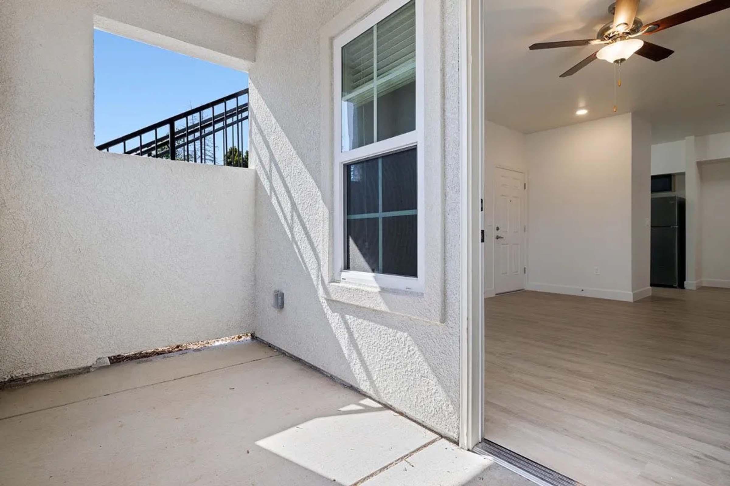 A corner of a room showing an open doorway leading to an outdoor space. The exterior features a textured white wall and a window. Inside, there’s a spacious area with light-colored flooring and a ceiling fan. Sunlight streams in, creating a bright atmosphere. A kitchen area is visible in the background.
