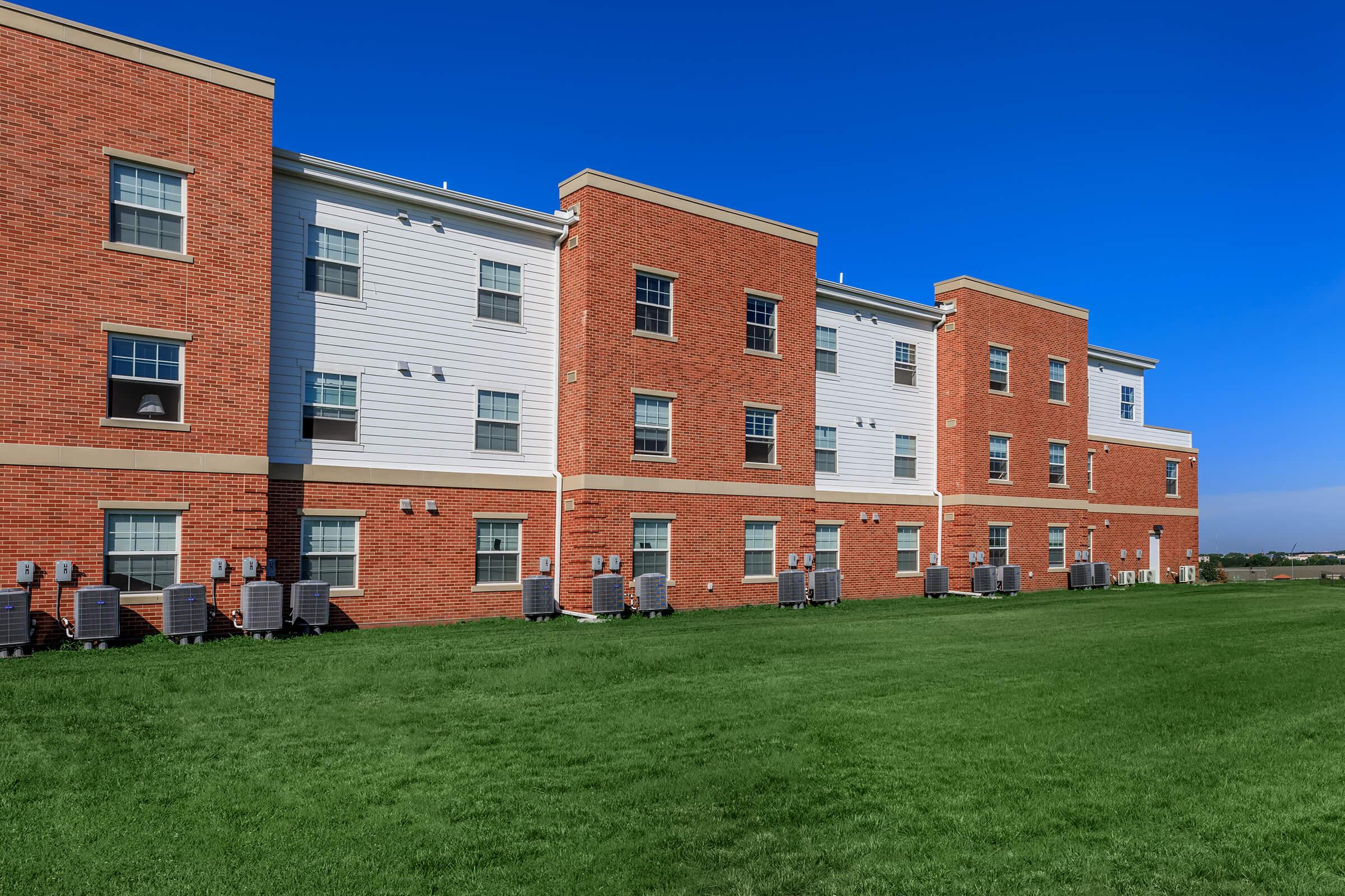 View of a multi-story red brick residential building with a white panel section, featuring several windows and air conditioning units along the base. The building is set against a clear blue sky, with a green lawn in the foreground.