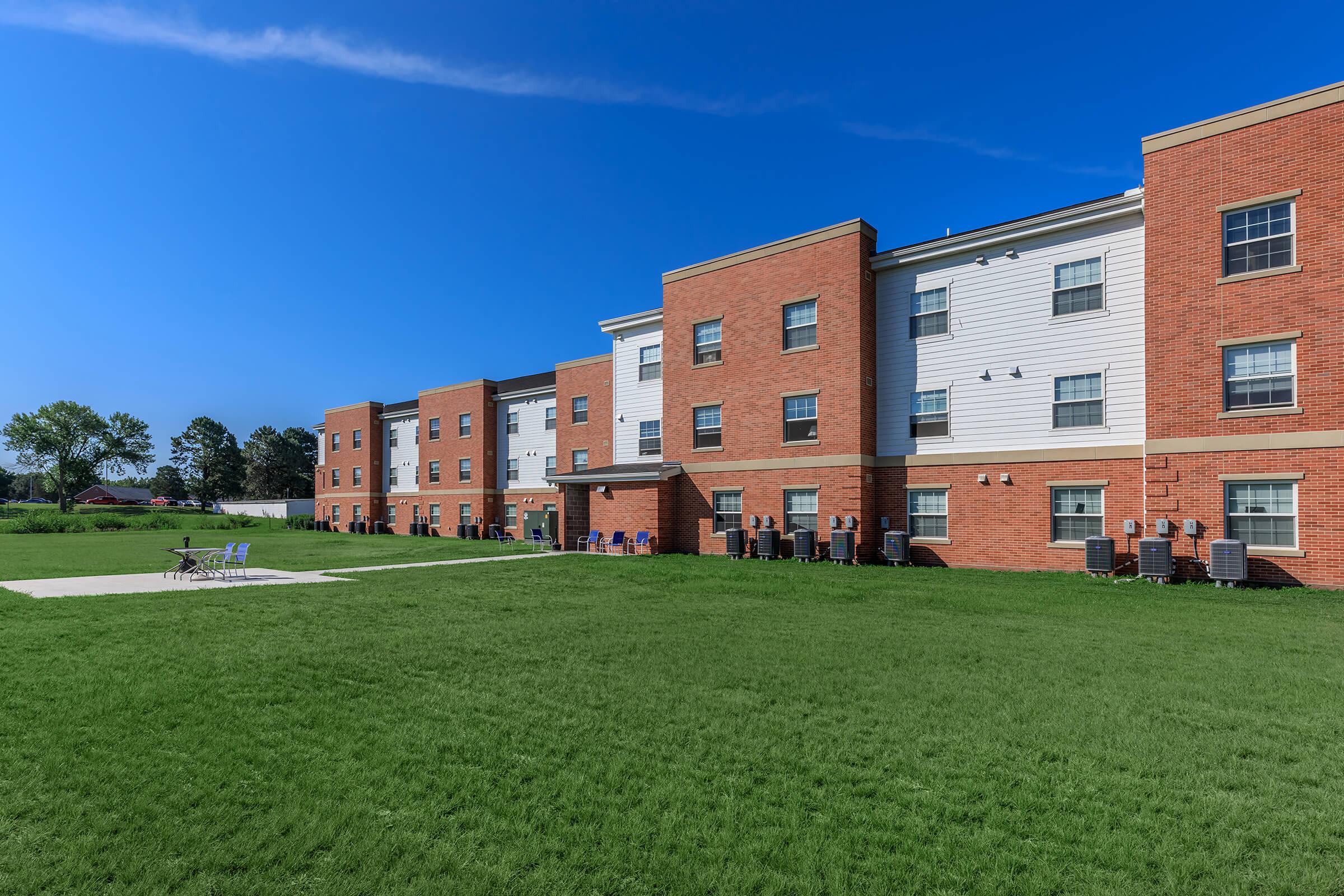 A brick apartment building with multiple windows and balconies, set against a clear blue sky. In the foreground, a grassy area features lounge chairs and a concrete patio. The scene is well-lit, highlighting the modern architecture and inviting outdoor space.