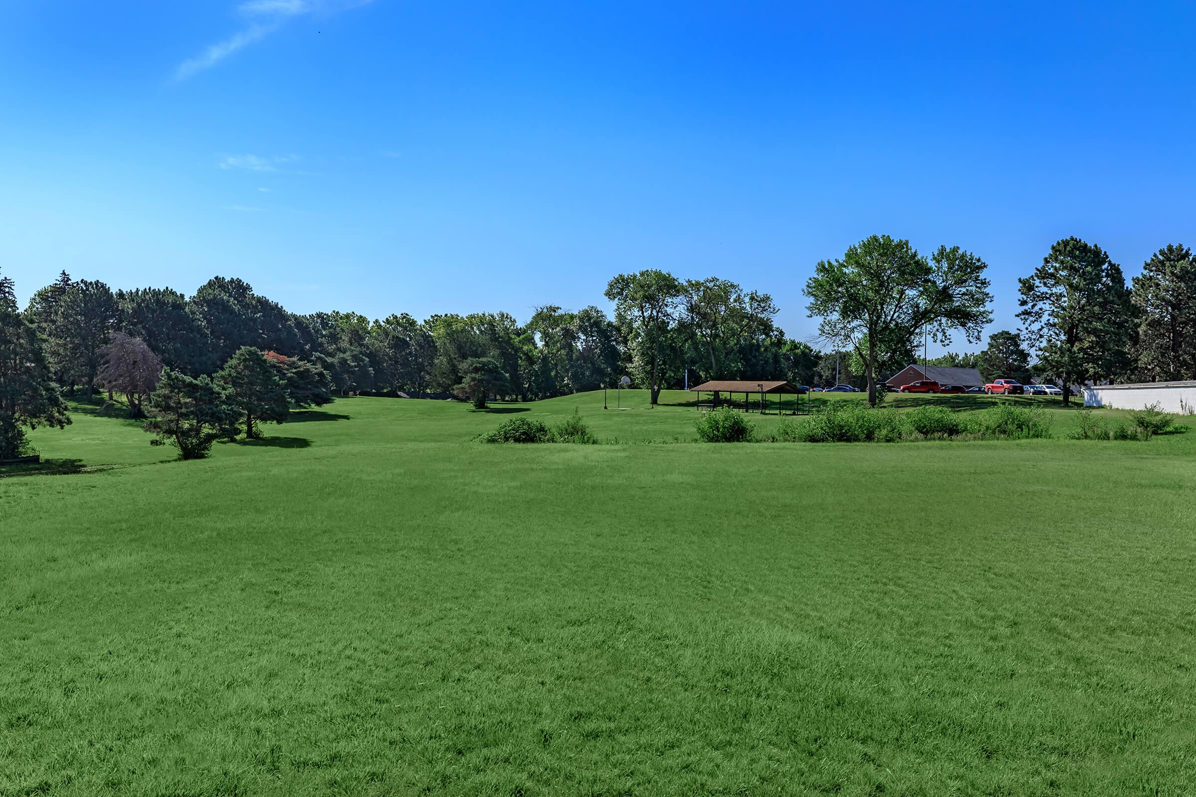 Lush green landscape under a clear blue sky, featuring rolling hills, scattered trees, and a distant view of a building and parked cars. The scene conveys a serene outdoor environment, ideal for recreational activities.