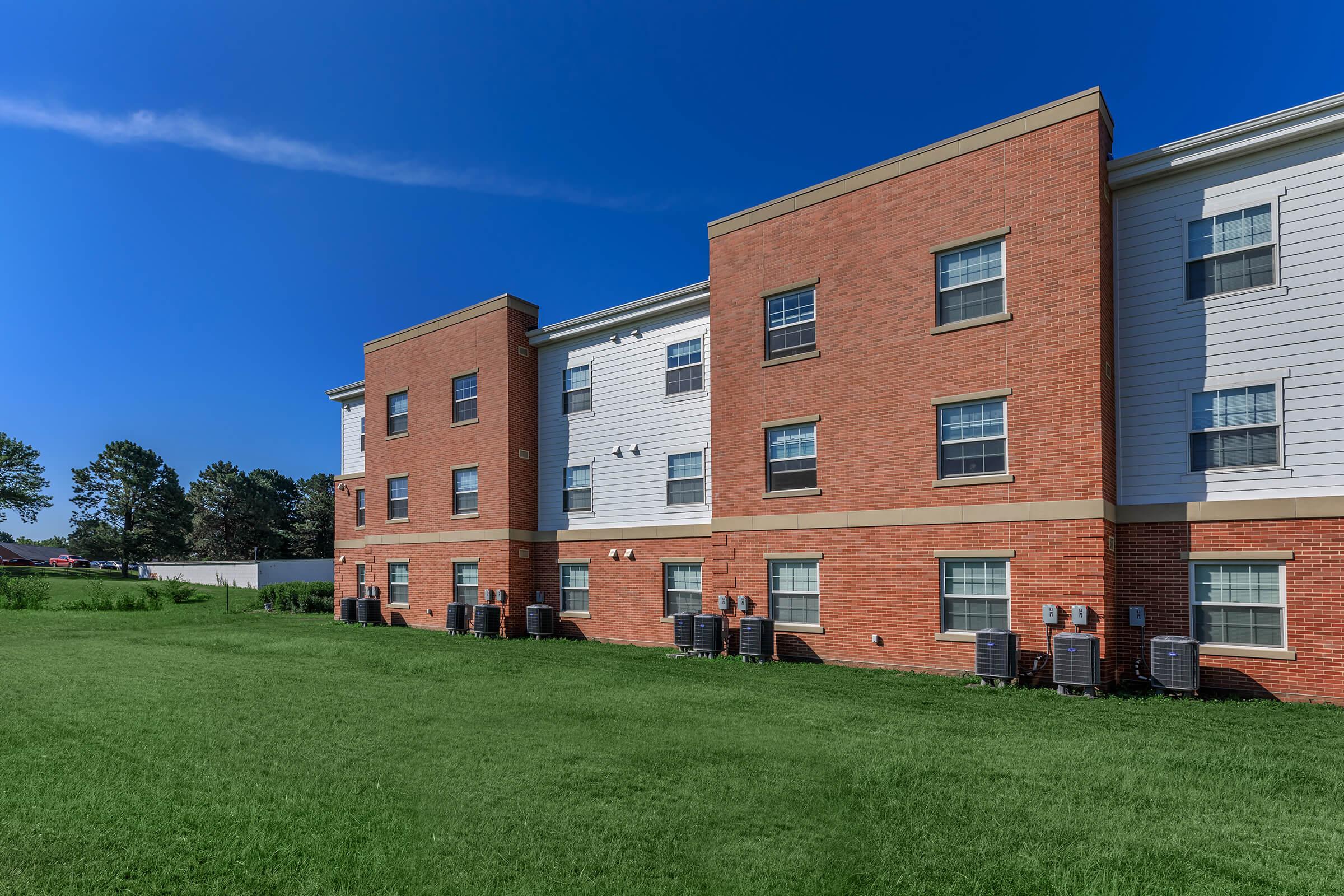 A two-story apartment building with a red brick facade and white siding. Several air conditioning units are visible on the ground floor. The building is surrounded by a green lawn and trees in the background, under a clear blue sky.