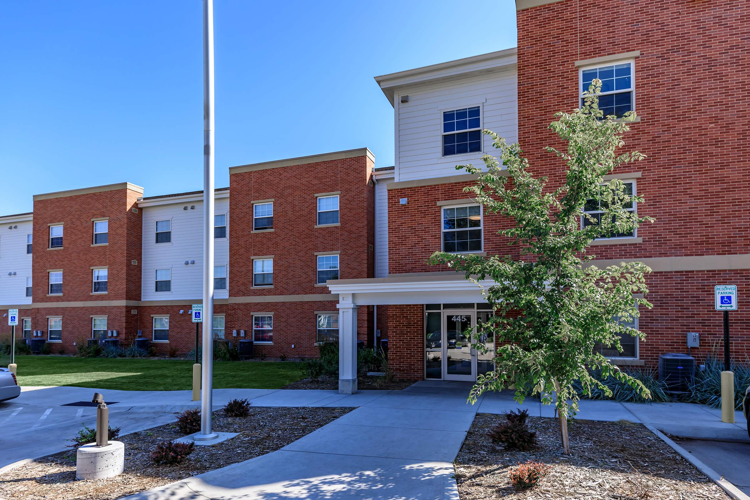 A modern brick building with multiple stories, featuring large windows and a landscaped entrance. The area includes a small garden with trees and shrubs, and there are accessible parking spaces nearby. Clear blue sky above enhances the welcoming atmosphere of the location.