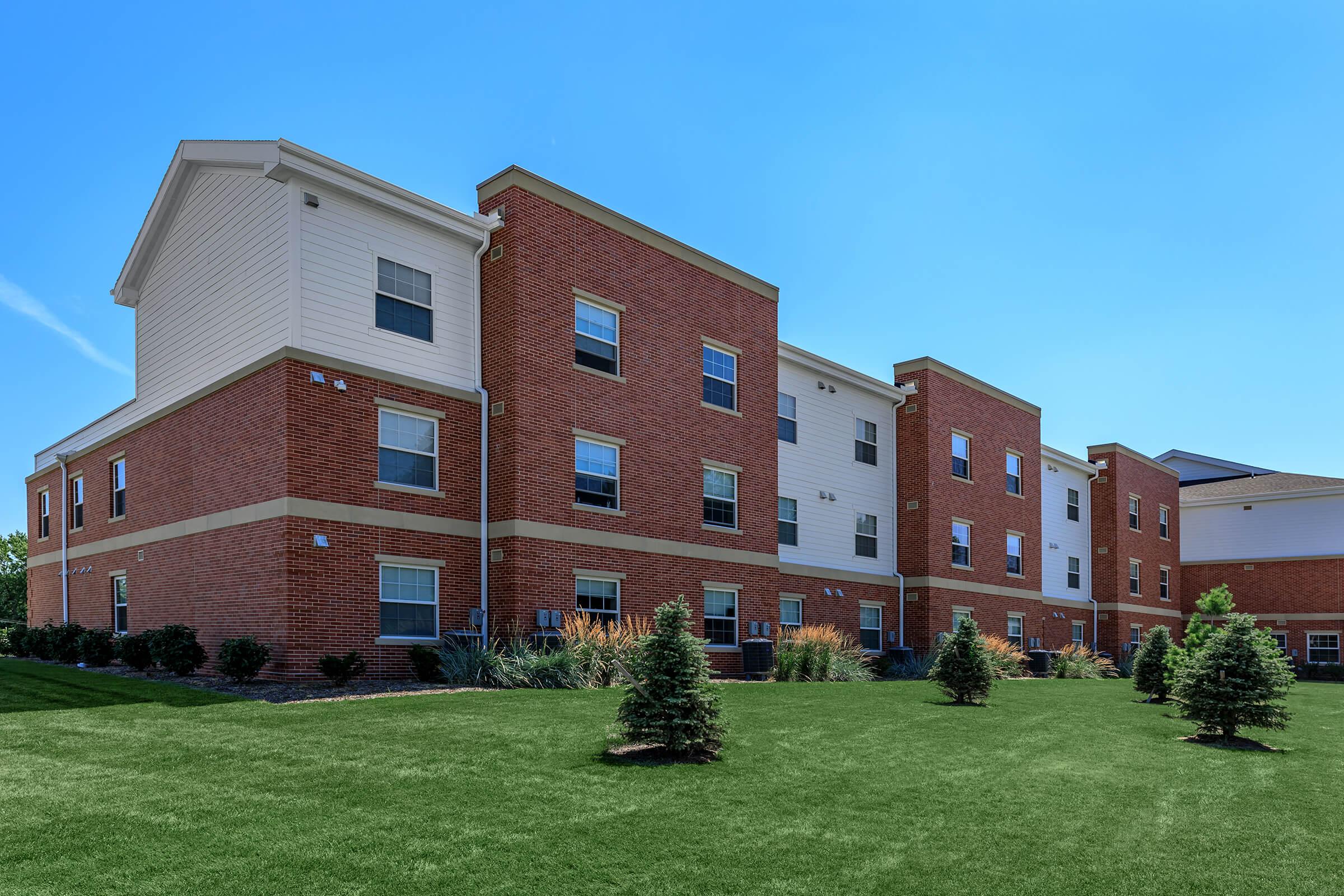 A modern brick apartment building with multiple stories, featuring large windows and a well-manicured lawn. The exterior includes a mix of brick and light-colored siding, with small evergreen shrubs planted in front on a sunny day. The sky is clear and blue, enhancing the vibrant appearance of the property.