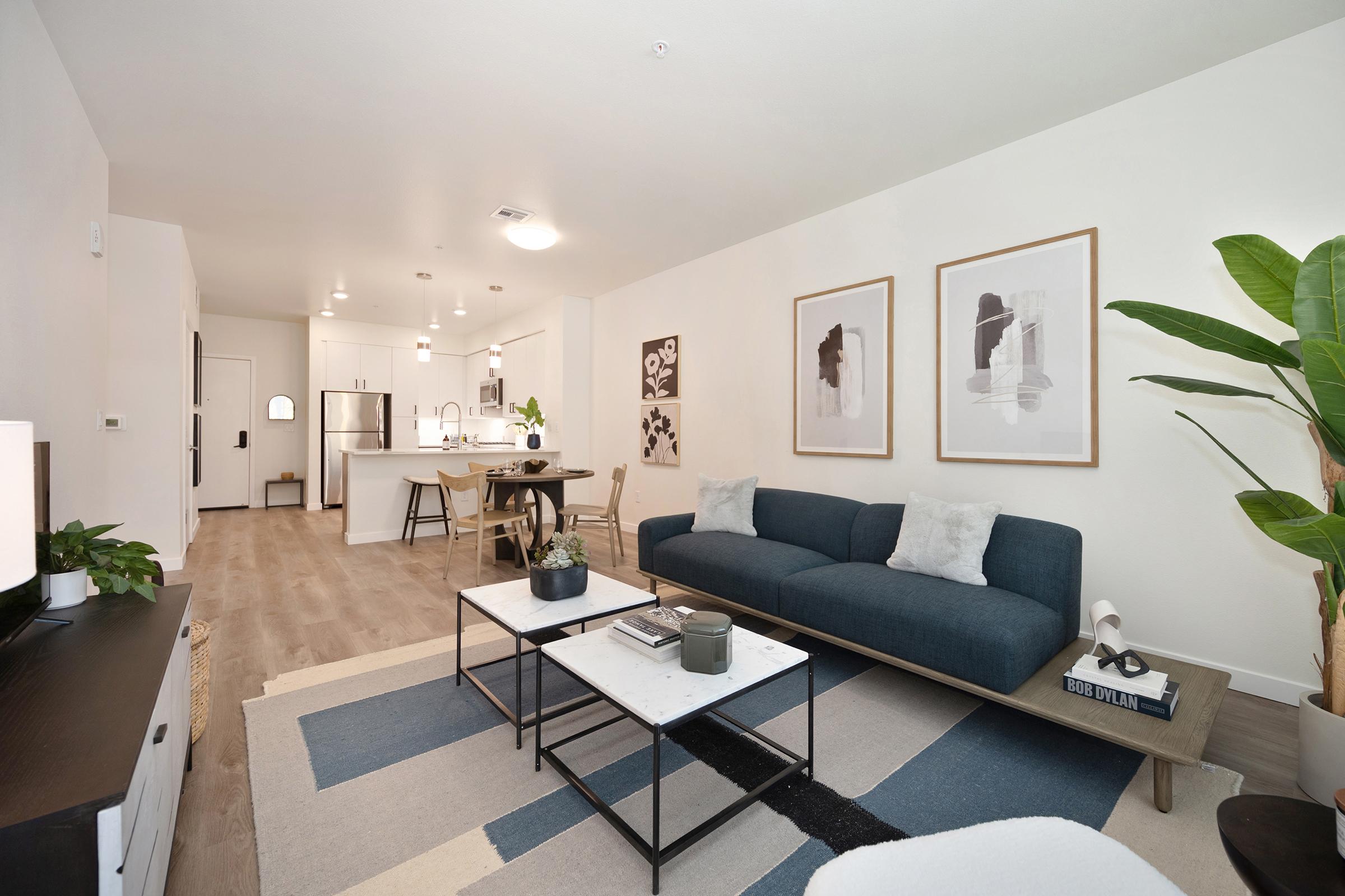A modern living room featuring a blue sofa, a coffee table with decorative items, and framed artwork on the walls. A dining area with wooden chairs and a kitchen with white cabinetry is visible in the background. Natural light enters through an open space, creating a bright and inviting atmosphere.