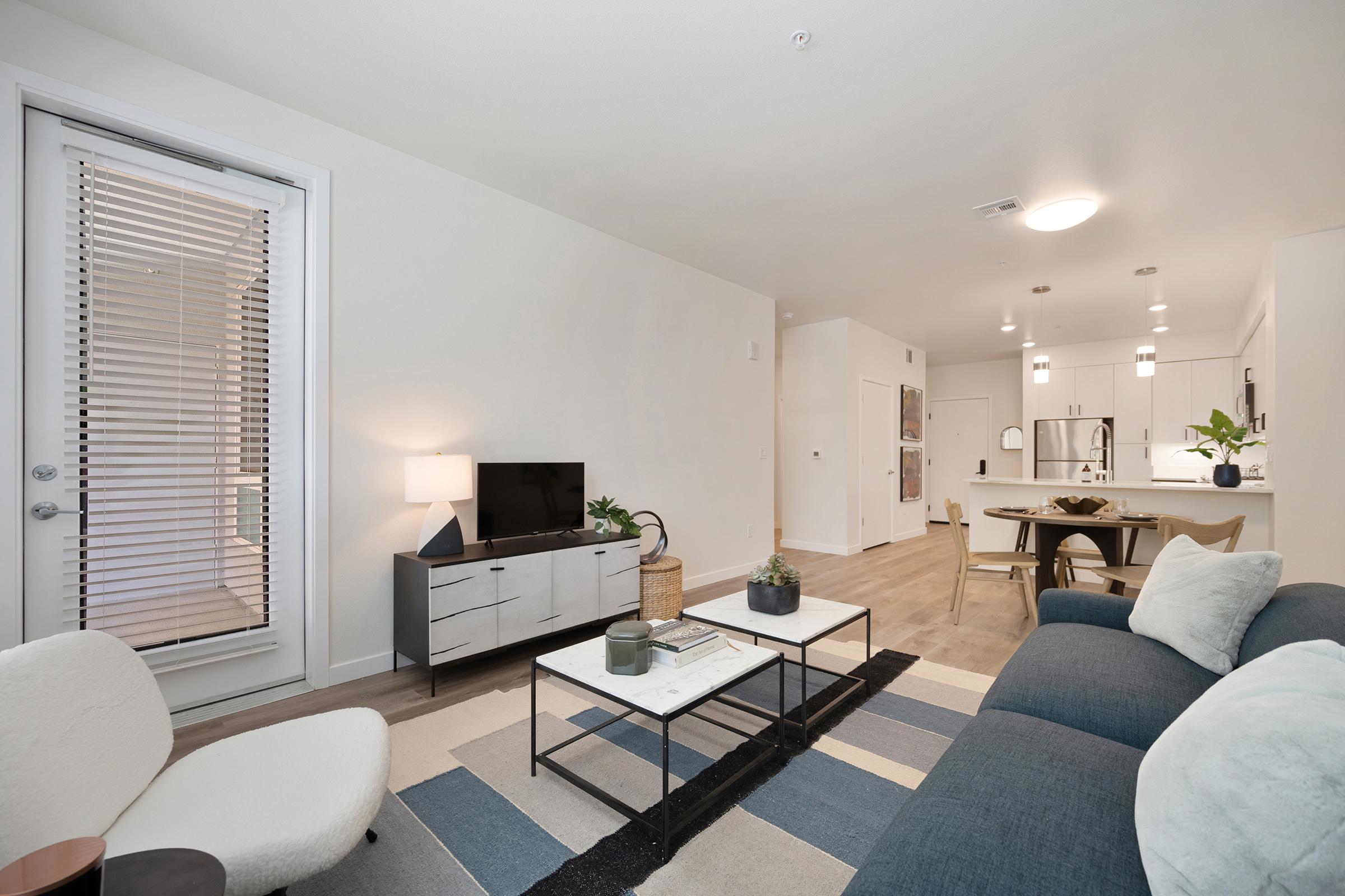 A modern living room featuring a comfortable blue sofa, a small round chair, and a coffee table on an area rug. There’s a flat-screen TV on a cabinet and a dining area visible in the background. Large windows with blinds allow natural light to fill the space, creating a bright and inviting atmosphere.