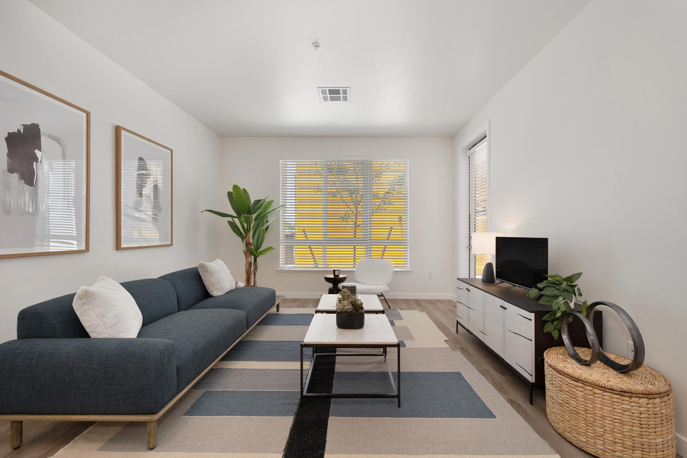 A modern, minimalist living room featuring a dark blue sofa, a stylish coffee table, and a white accent chair. Large windows allow natural light, showcasing a green plant and contemporary artwork on the walls. A decorative basket and a TV stand add functionality to the space.