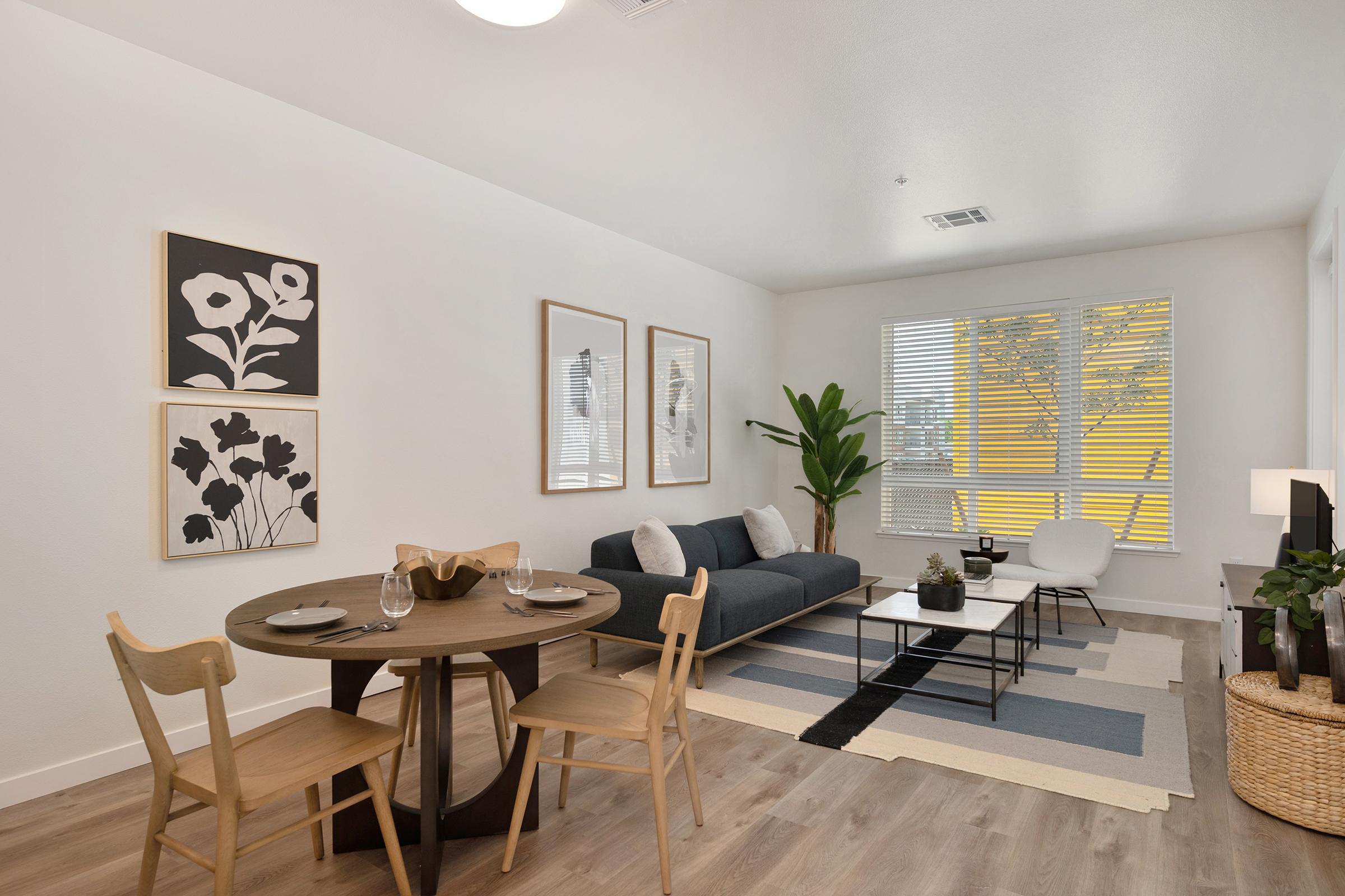 A modern living room featuring a grey sofa with decorative cushions, a round dining table set for two, and stylish artwork on the walls. There is a potted plant in the corner, a light rug on the floor, and large windows allowing natural light to fill the space.