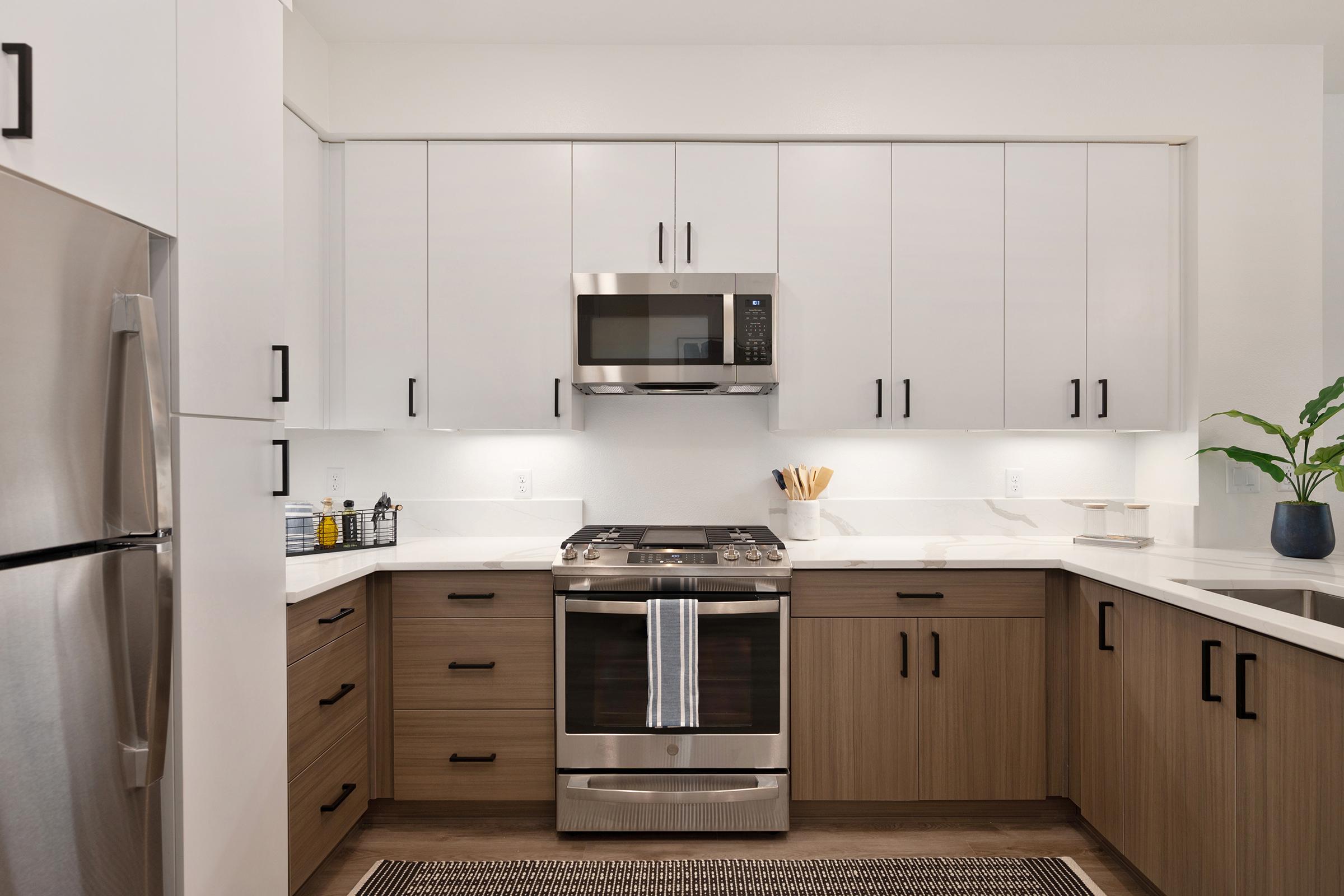 Modern kitchen featuring sleek cabinetry, stainless steel appliances, a gas range, and a microwave. The design combines white upper cabinets with wood lower cabinets, complemented by a light countertop. A small potted plant and kitchen utensils add a touch of greenery and functionality to the space.
