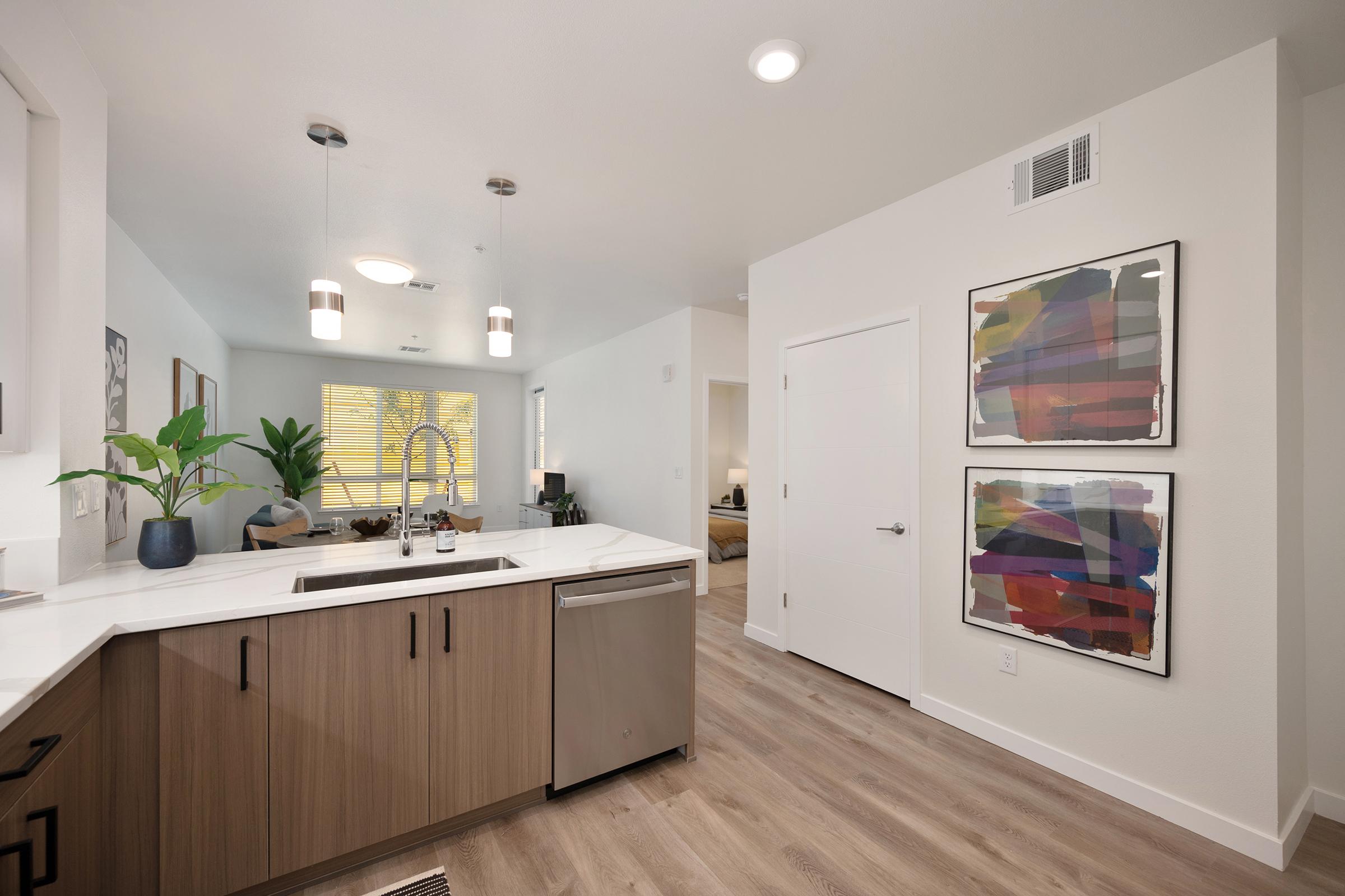 Modern kitchen with light wood cabinets and a stainless steel sink. Pendant lights hang above the kitchen island. A green plant is placed near the window, and colorful abstract art is framed on the wall. The open layout leads to a cozy living area visible in the background.