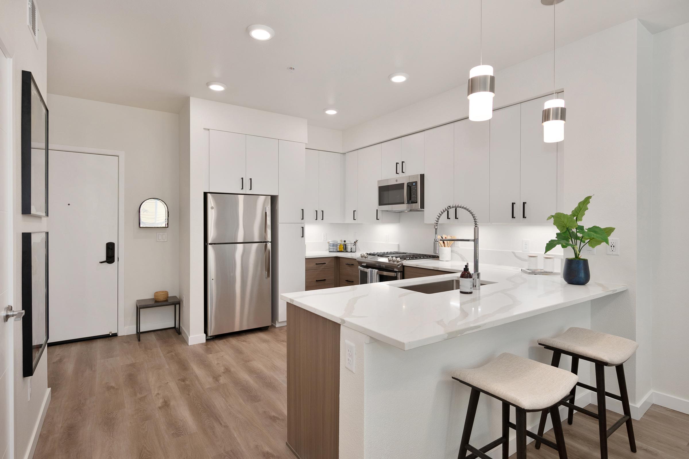 A modern kitchen featuring sleek white cabinetry, stainless steel appliances, and a large island with seating. The countertops are white, complemented by hardwood flooring. The space is bright and spacious, with recessed lighting and a potted plant for decoration. A door to the left leads outside.