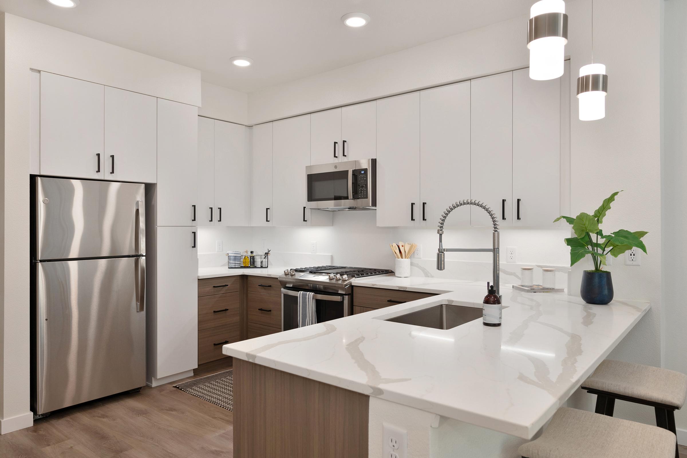 Modern kitchen featuring white cabinets, stainless steel appliances, and a marble countertop. The layout includes a refrigerator, microwave, and gas stove. A plant sits on the counter along with kitchen utensils, and there are stylish pendant lights overhead, creating a bright and inviting atmosphere.