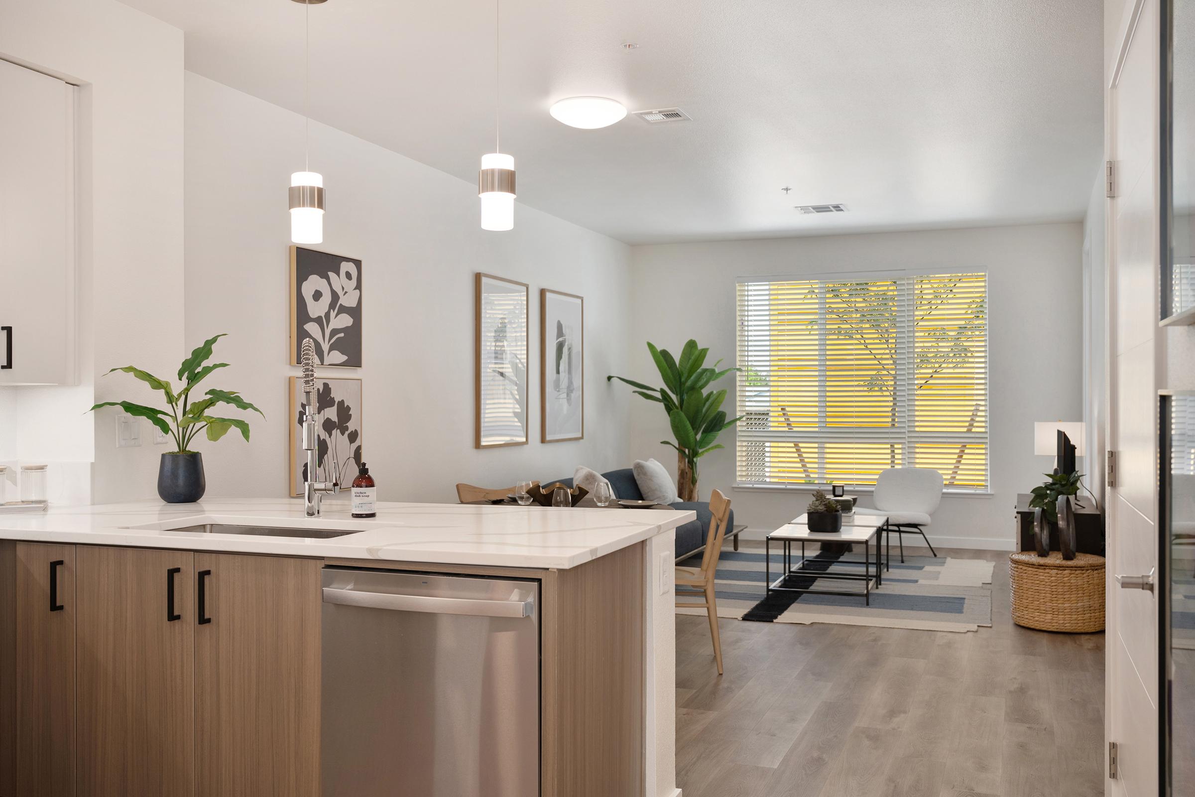 Modern kitchen and living area featuring a sleek countertop, stainless steel appliances, and a dining space. The living room includes stylish furniture, plants, and large windows allowing natural light. Neutral tones and contemporary decor create a cozy atmosphere.