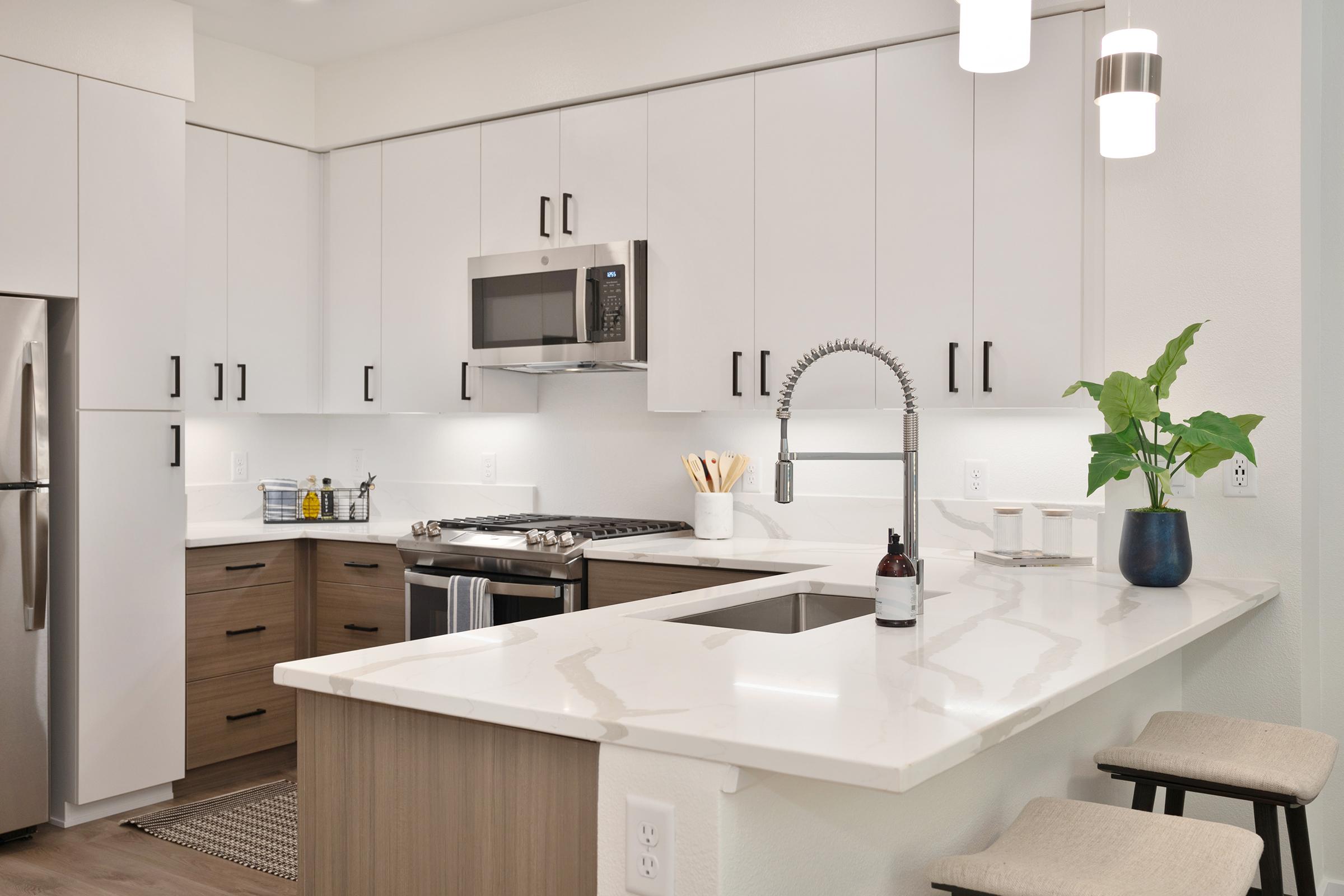 Modern kitchen featuring sleek white cabinets, stainless steel appliances, a gas stove, and a large island with a countertop. The space includes a decorative plant and minimalist bar stools, enhancing the contemporary design with warm wood accents and soft lighting.