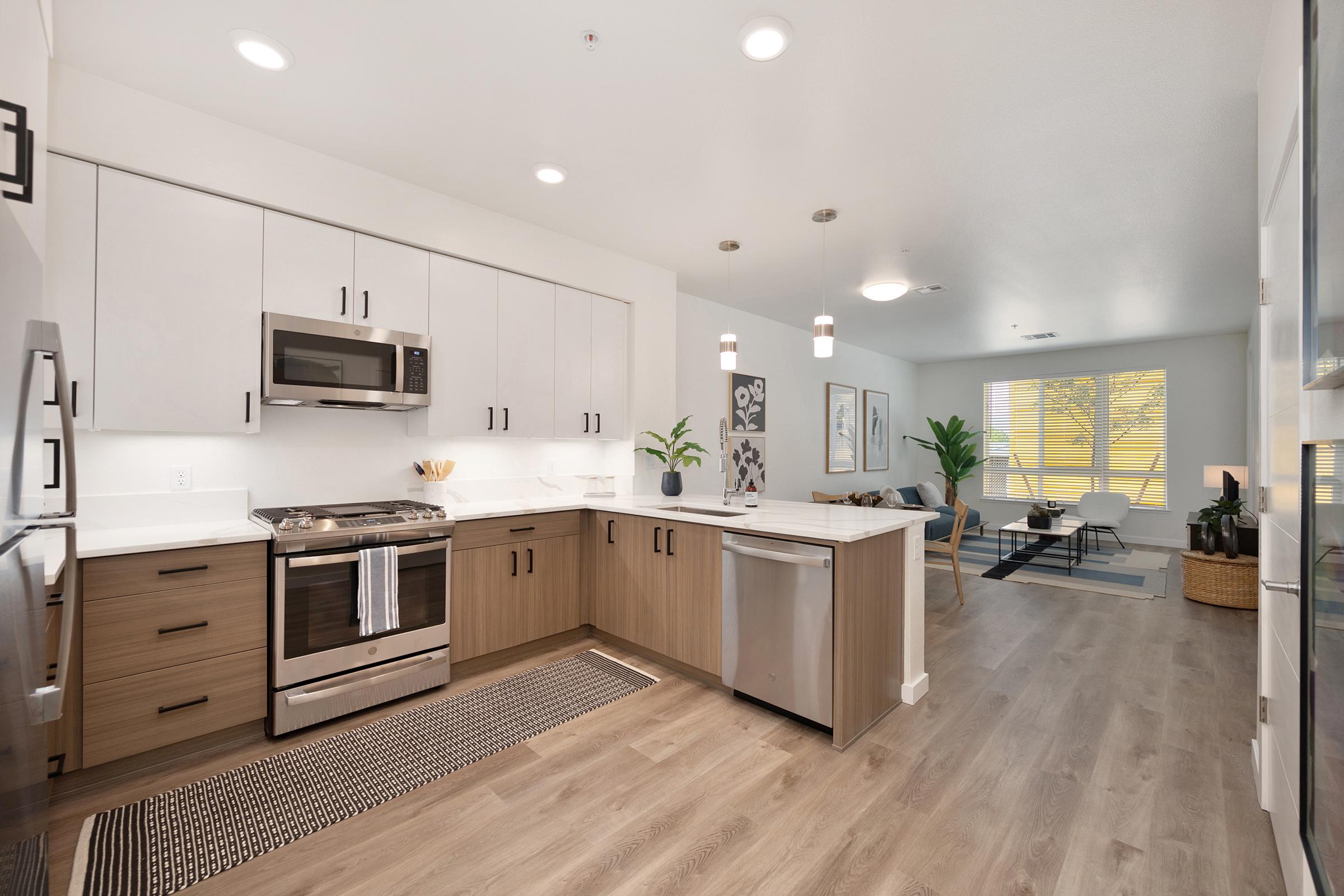 Modern kitchen with stainless steel appliances, including a microwave and oven. The cabinetry is a mix of white and light wood, complemented by a stylish backsplash. A cozy living area is visible in the background, featuring seating and large windows with natural light. The floor has a warm wood finish.