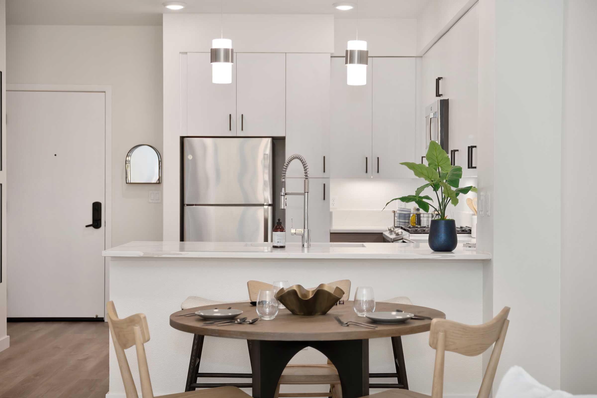 A modern kitchen and dining area featuring a round table set with plates and glassware, surrounded by wooden chairs. The kitchen showcases stainless steel appliances, white cabinetry, and a decorative plant on the countertop. Bright lighting enhances the open and contemporary atmosphere.