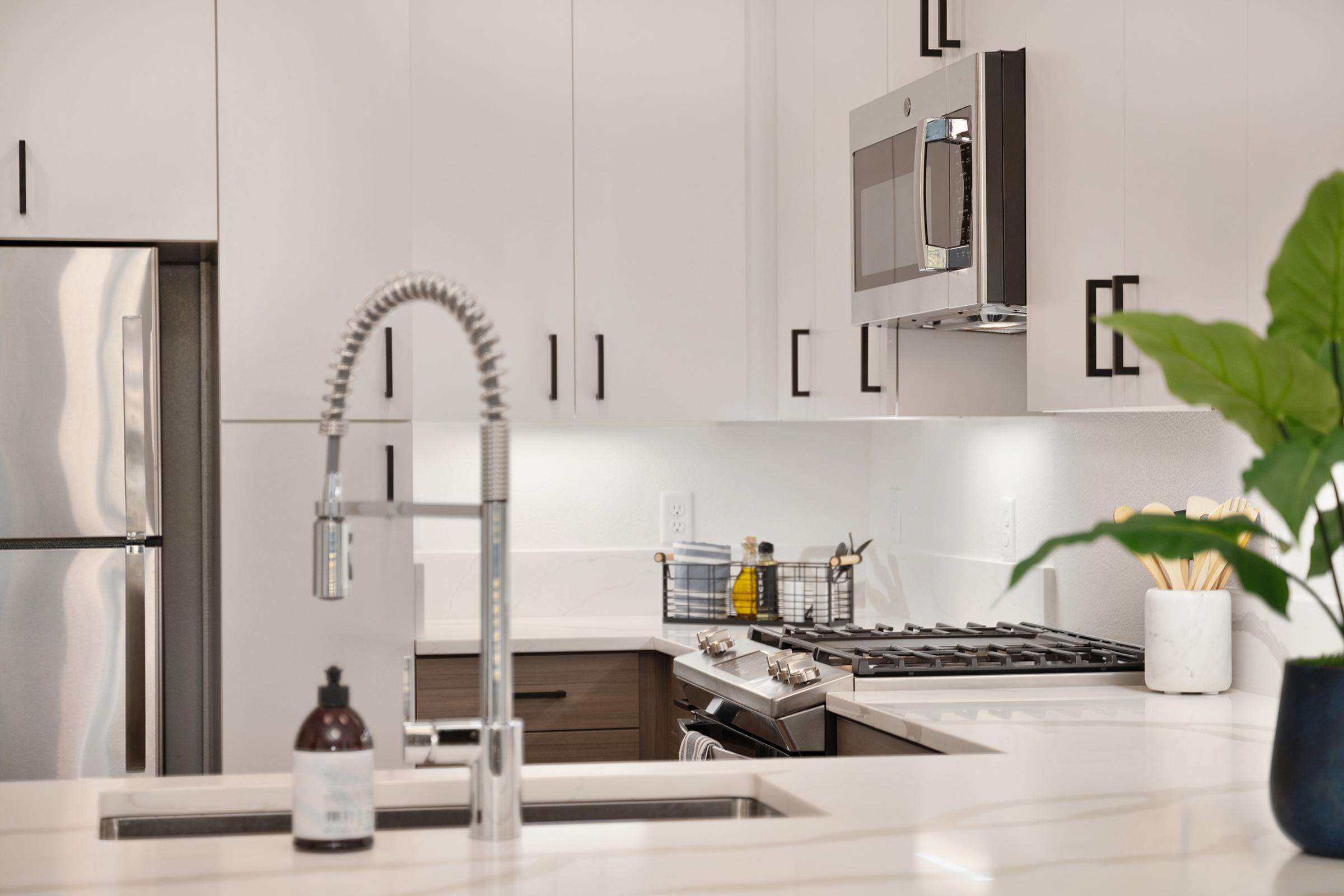 Modern kitchen featuring sleek white cabinetry, stainless steel appliances, and a gas stove. A contemporary faucet is installed over a double sink, with a decorative plant and kitchen essentials visible on the counter. The design showcases a clean and inviting atmosphere with bright lighting.
