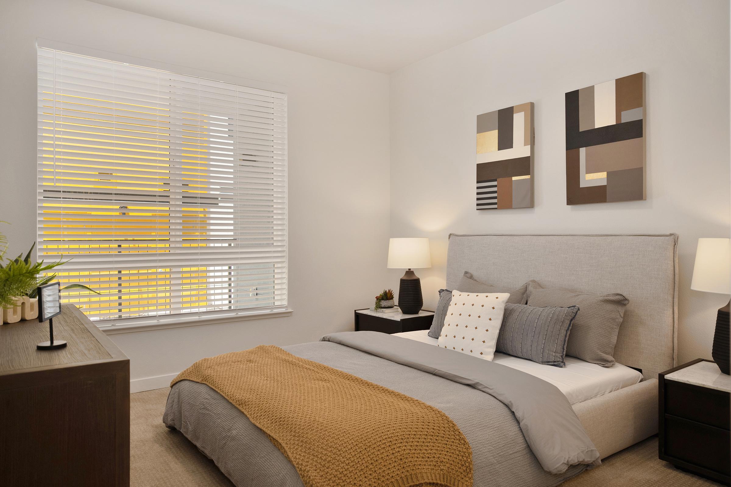 A cozy bedroom featuring a light-colored bed with a gray blanket and decorative pillows. There are two modern abstract paintings on the wall, bedside tables with lamps, and a dresser. Natural light filters through a window with white blinds, illuminating the space.