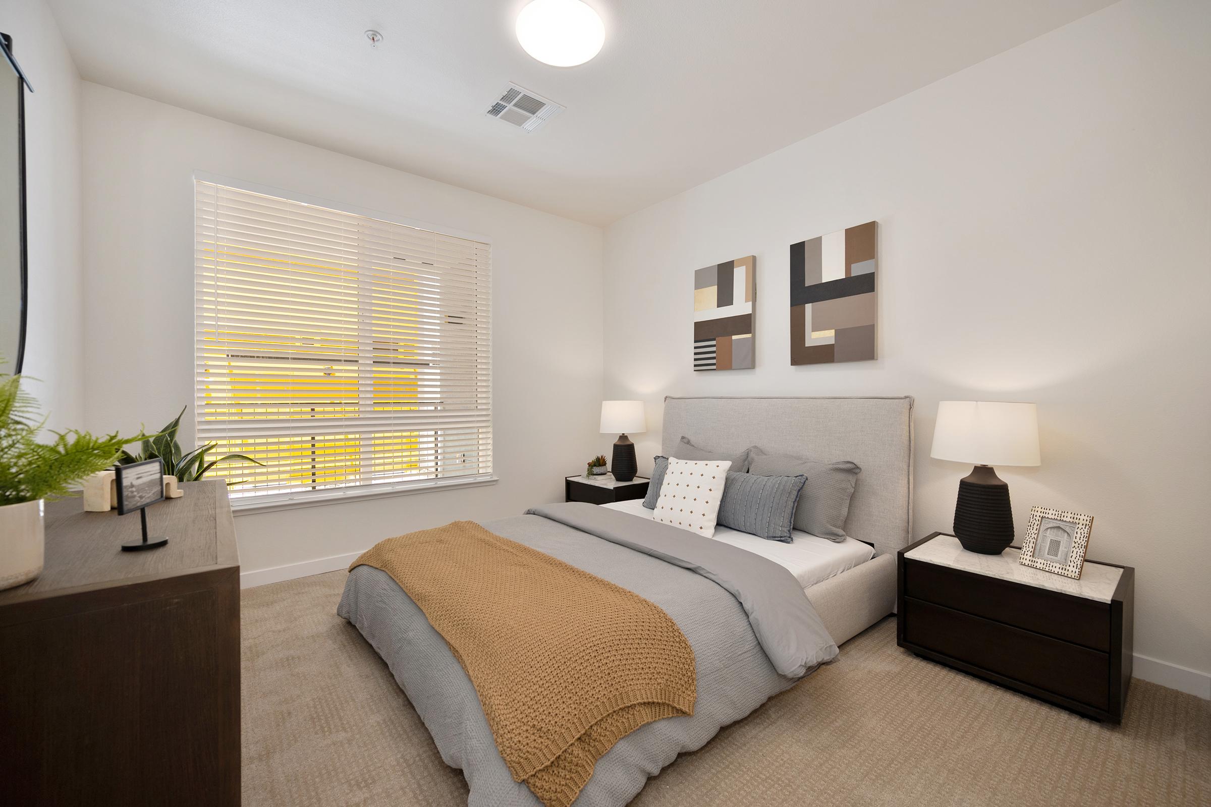 A cozy bedroom featuring a queen-sized bed with gray bedding and a brown throw. There are two nightstands with lamps on either side, and a minimalist dresser. The room has large windows with blinds, allowing natural light to fill the space, and abstract artwork is displayed on the wall.