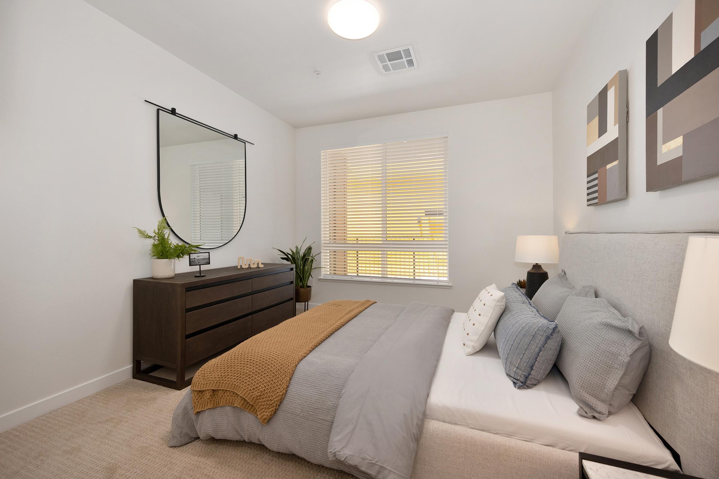 A modern bedroom featuring a neatly made bed with gray bedding and decorative pillows, a stylish round mirror above a dark wood dresser, and a small plant on the dresser. Natural light flows through a window with blinds, enhancing the cozy atmosphere of the room.