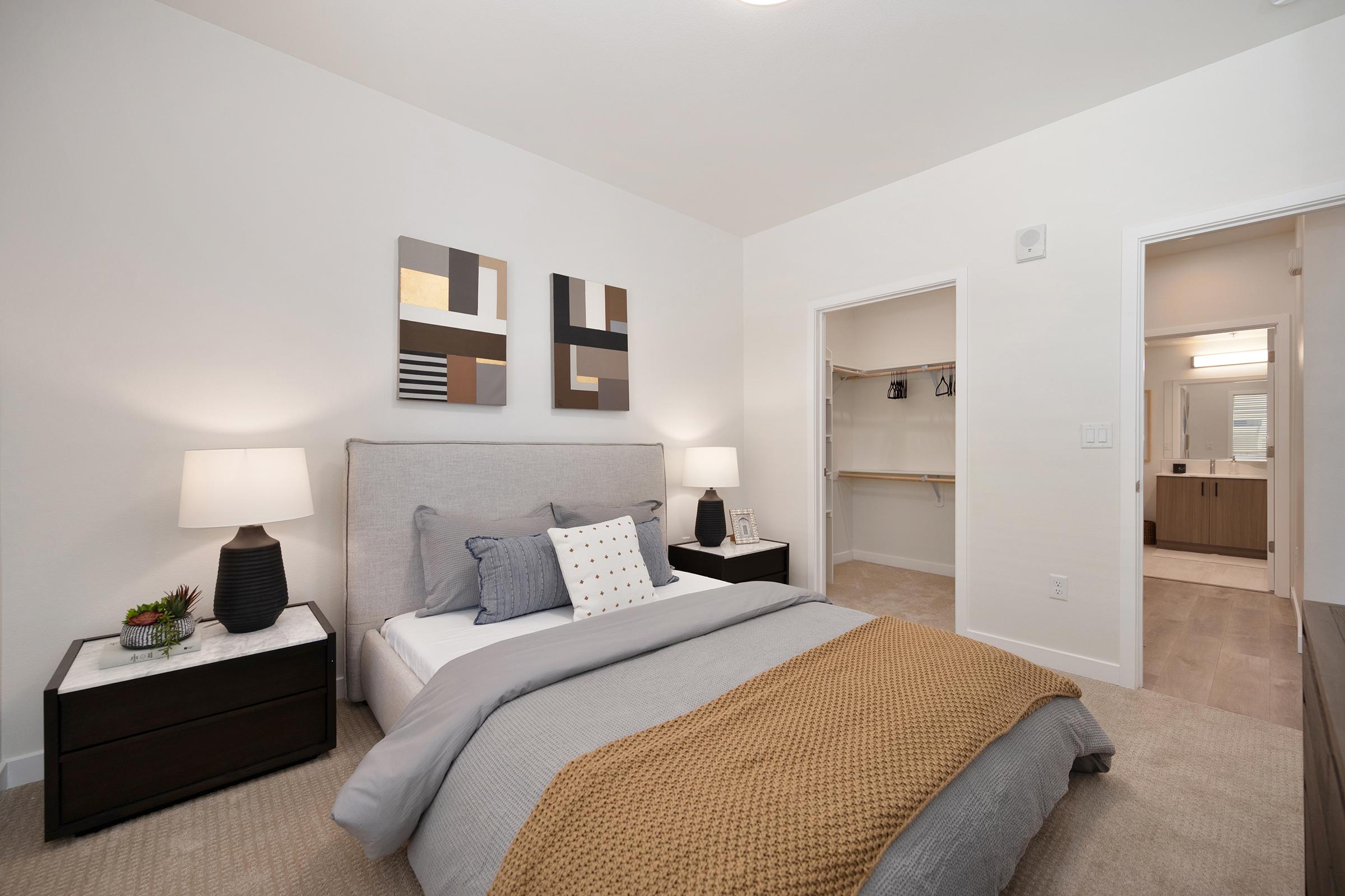 A cozy bedroom featuring a queen-sized bed with a light gray blanket and decorative pillows. Two modern lamps sit on black nightstands. The walls are adorned with abstract artwork. An open closet space is visible on the right, leading to a bathroom area with neutral tones, creating a warm and inviting atmosphere.