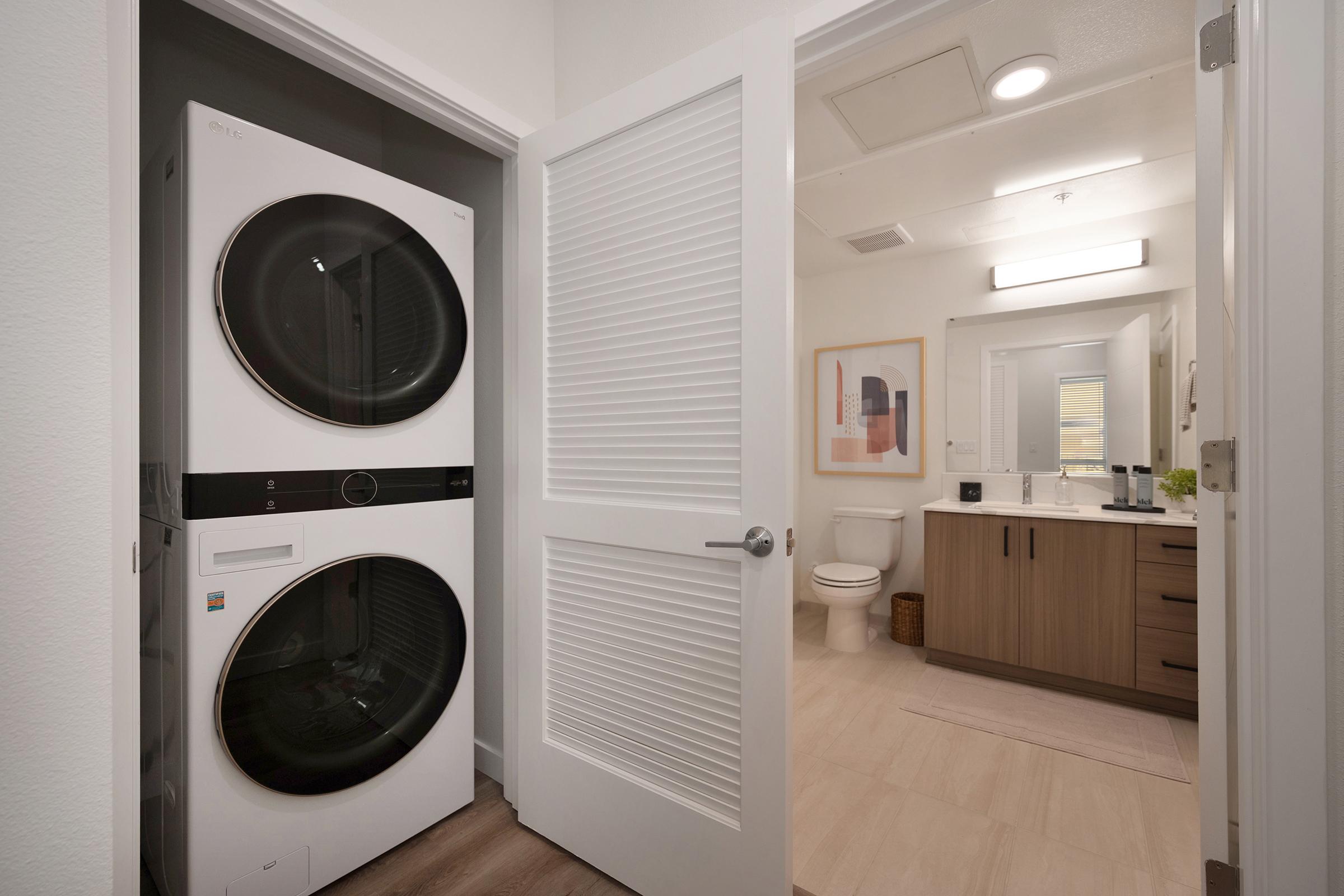 A modern laundry area with a stacked washer and dryer located in a bathroom entrance. The bathroom features a light wood vanity, a toilet, and a mirror with soft lighting. The walls are painted in neutral tones, and there is a framed artwork on the wall, contributing to a clean and stylish design.