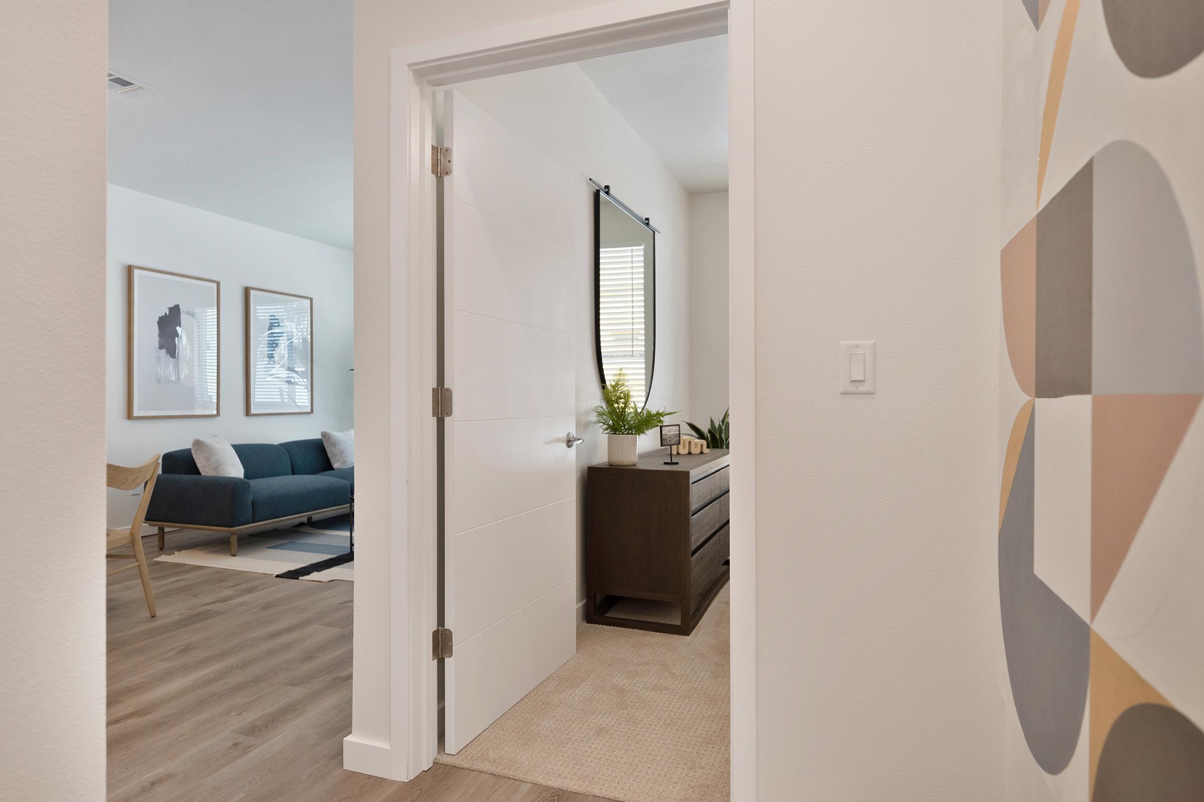 A view from a hallway into a stylish living room featuring a blue sofa and framed artwork. A doorway leads to a well-decorated room with a dresser and plants, creating a modern and inviting atmosphere. Light streams in through the window, enhancing the warm, neutral color palette.