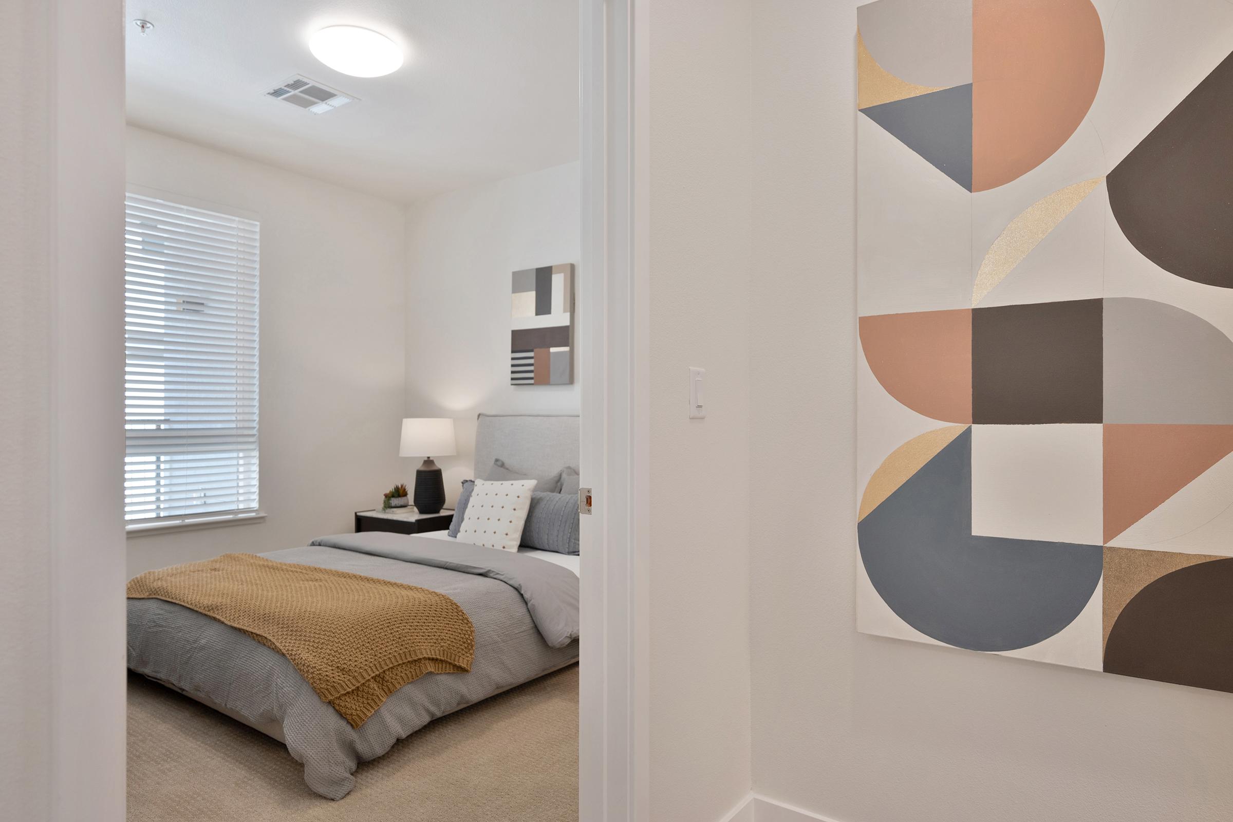 A view from a doorway into a modern bedroom featuring a neatly made bed with gray linens and a decorative yellow throw. A bedside lamp is on a nightstand, and a large abstract painting with geometric shapes in earthy tones is visible on the wall. Soft natural light enters through a window with white blinds.