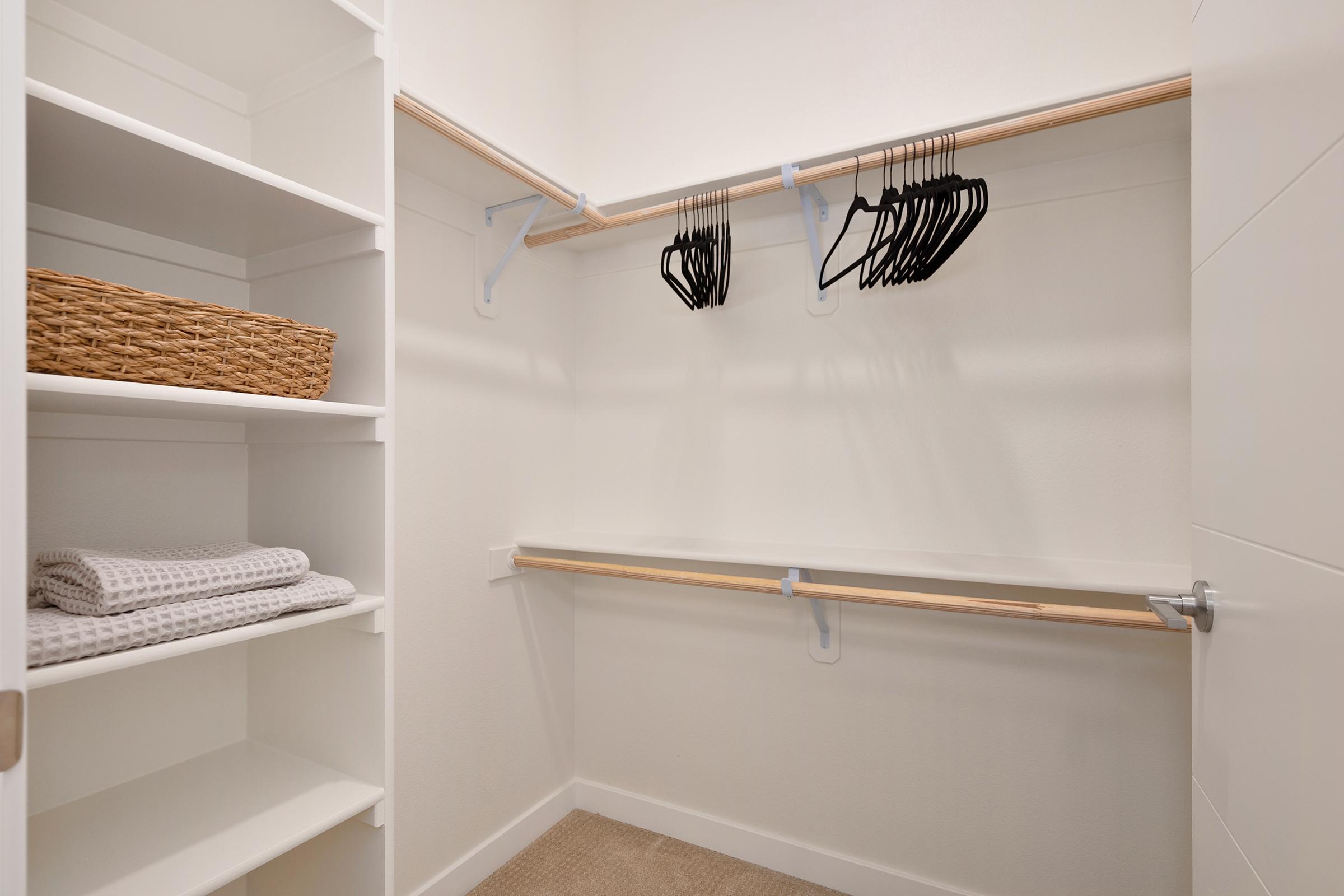 A clean and organized closet featuring white shelves, a wooden hanging rod with black hangers, a basket on the shelf, and neatly folded towels on the lower shelf. The walls are a light color, and the flooring is carpeted, creating a bright and airy atmosphere.