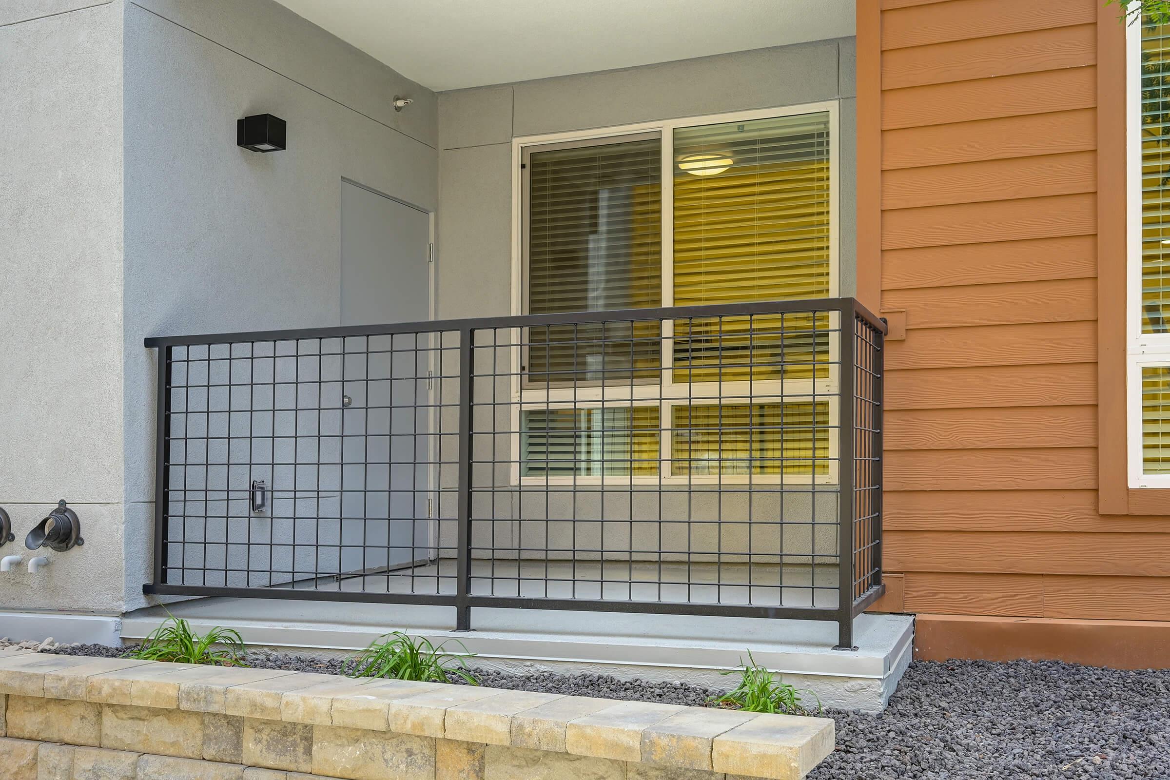 A modern apartment balcony with a metal fence, featuring a door to the interior and large windows letting in natural light. The floor is a smooth concrete surface, and there are small plants along the edge. The building has a contrasting exterior with both grey and brown siding.