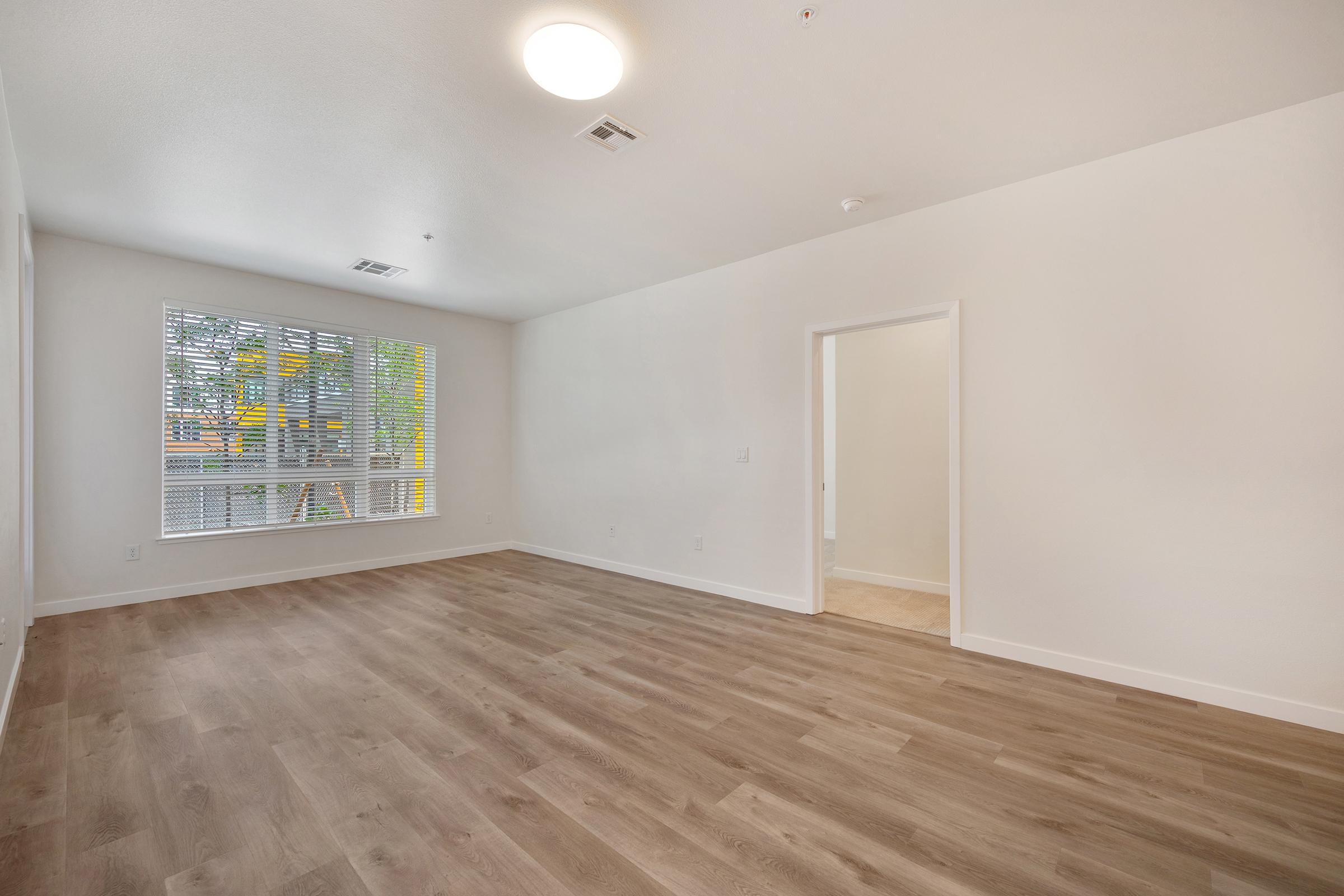A spacious and empty room featuring light-colored walls and a wood-style floor. There is a large window with blinds, allowing natural light to enter. A door on the right leads to another space, while the ceiling has a round ceiling light fixture. The overall atmosphere is bright and minimalist.