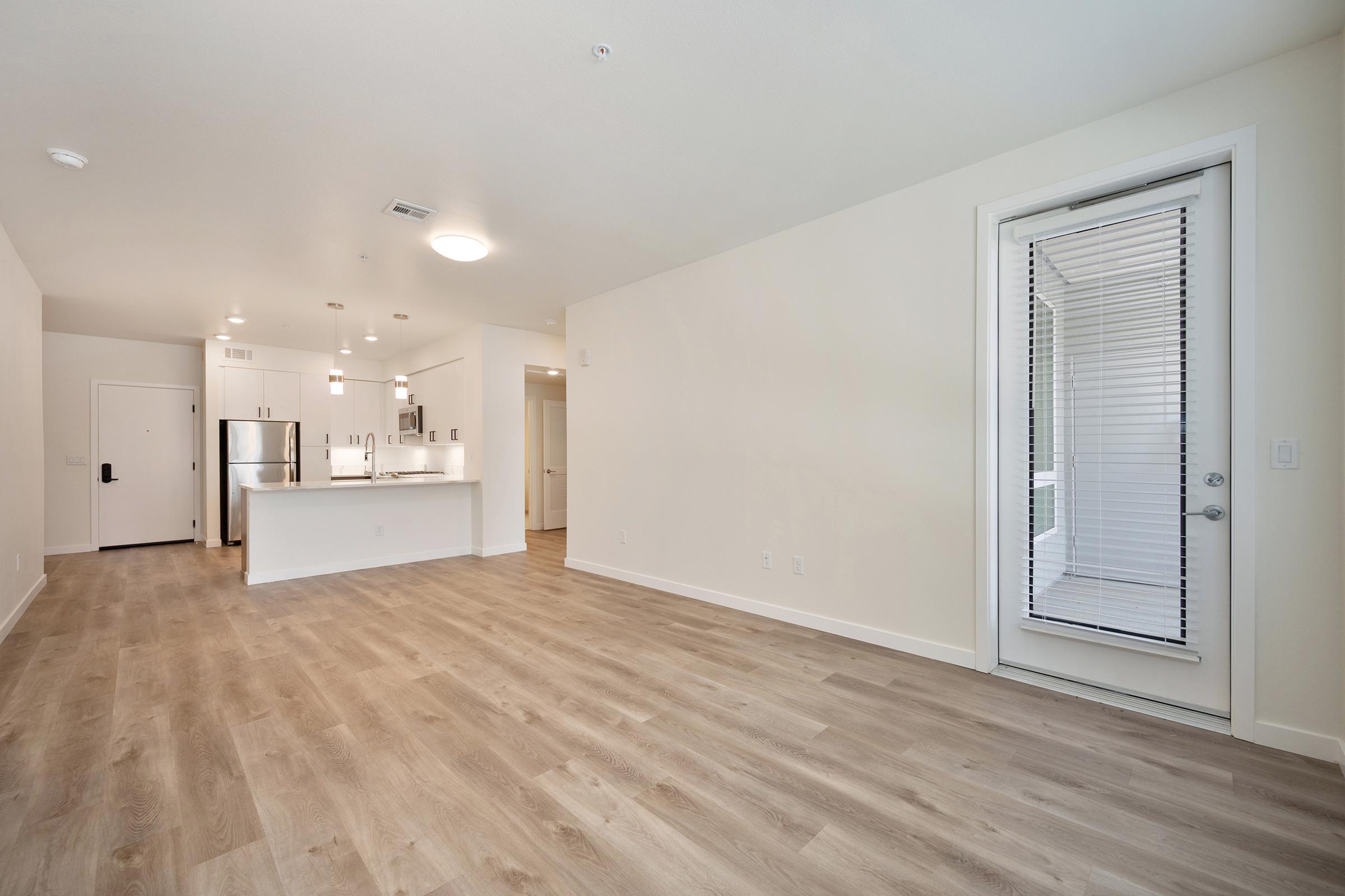 A spacious, modern interior of an empty apartment featuring light wood flooring, a white kitchen with stainless steel appliances, and a door leading to an outdoor area. The walls are painted in neutral tones, and there is ample natural light in the room.